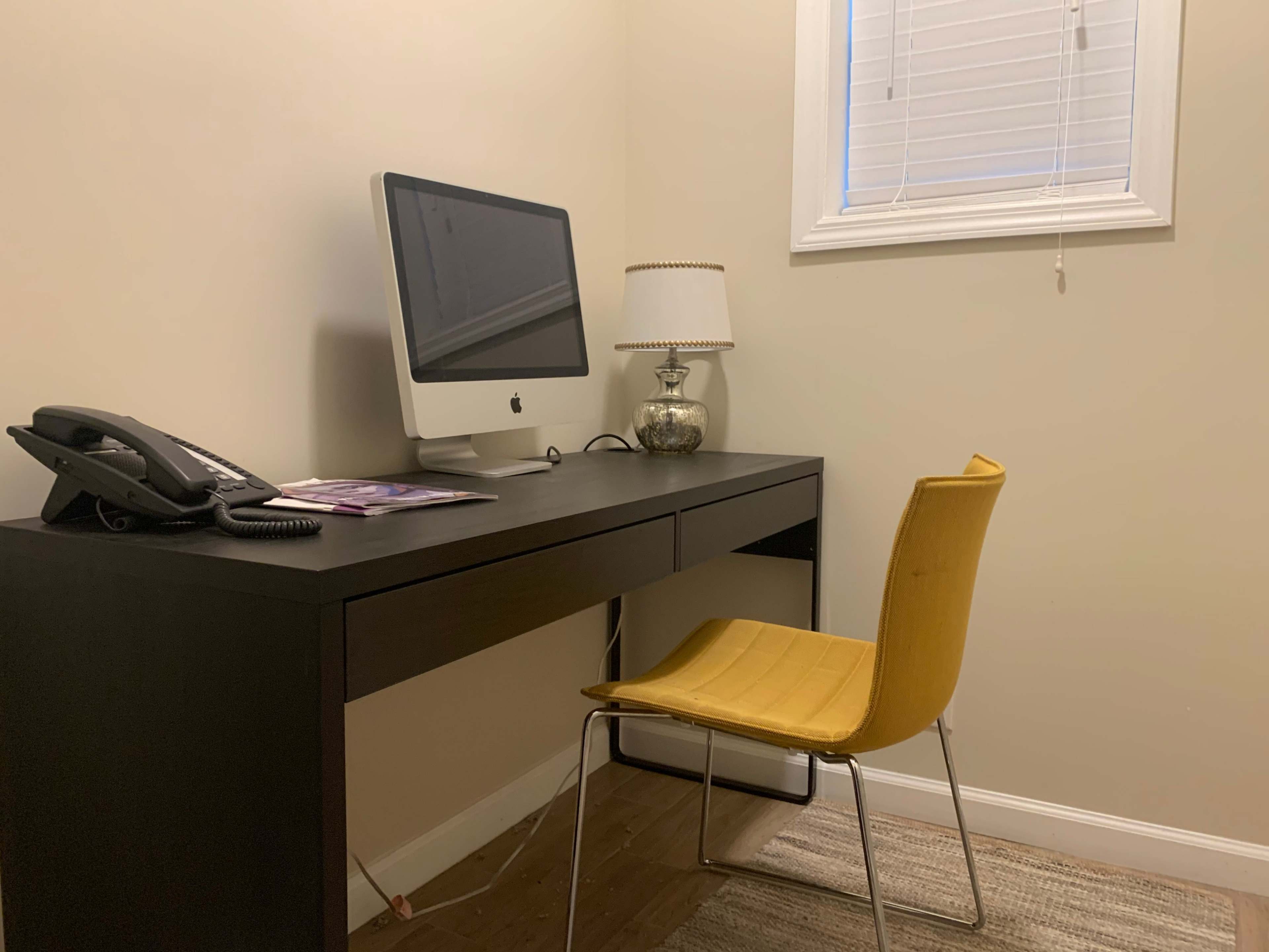 A simple workspace features an iMac on a black desk, a yellow chair, a telephone, and a lamp beside a window with closed blinds.