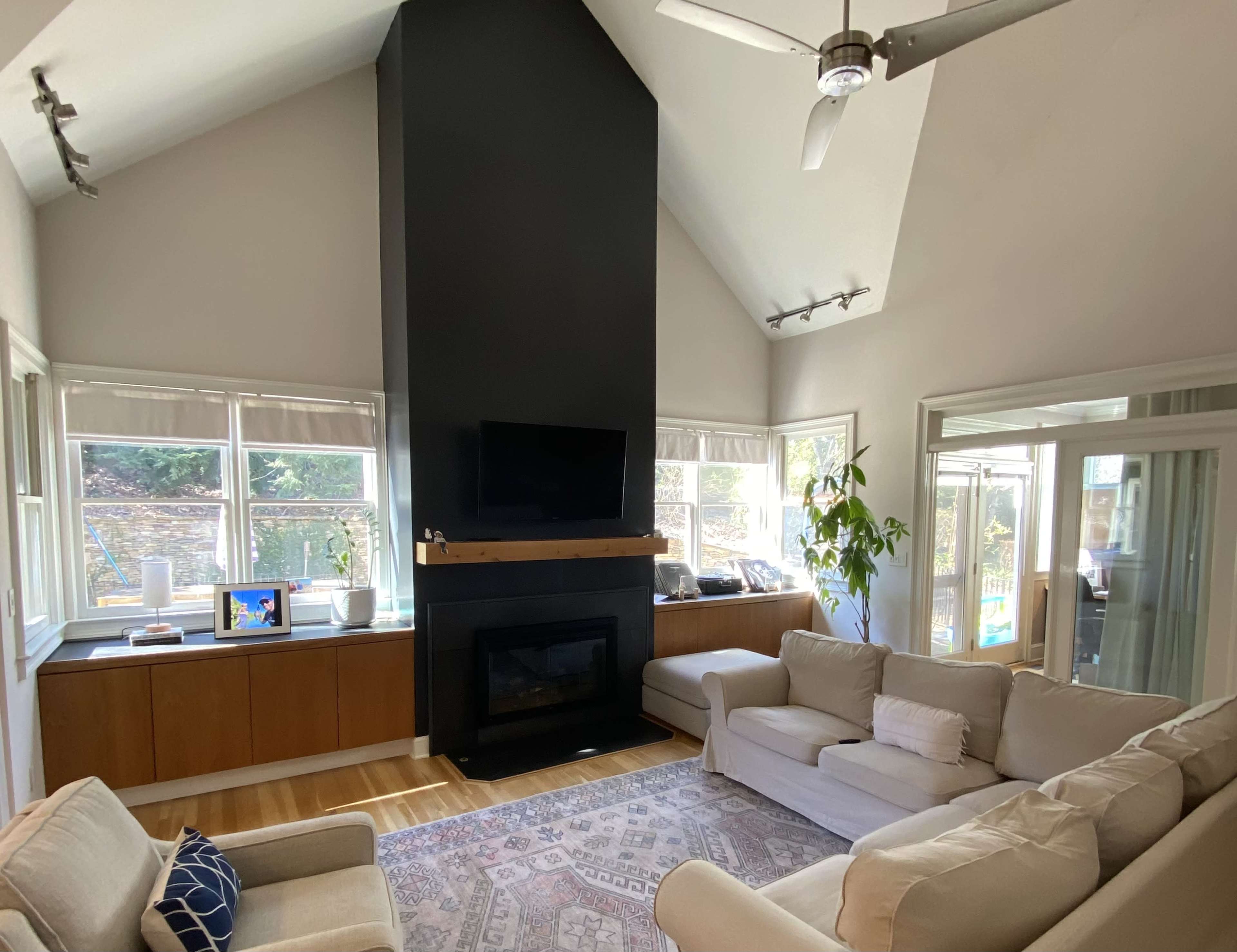 The image shows a living room with a high ceiling, featuring a black fireplace, a television mounted on the wall, large windows, and a comfortable seating area.