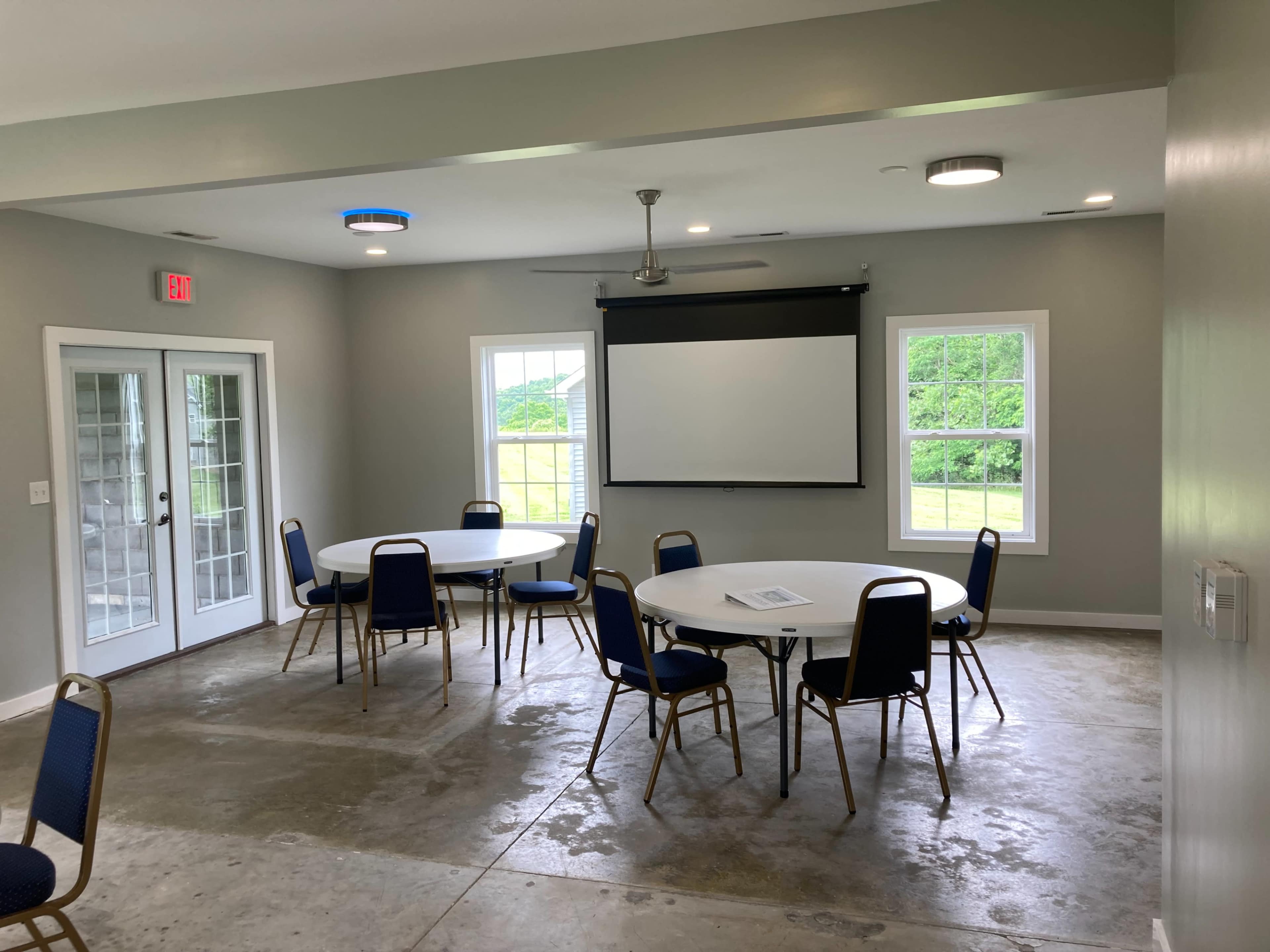 A well-lit meeting room features several round tables with chairs, a retractable screen, and double doors leading to an outdoor view.