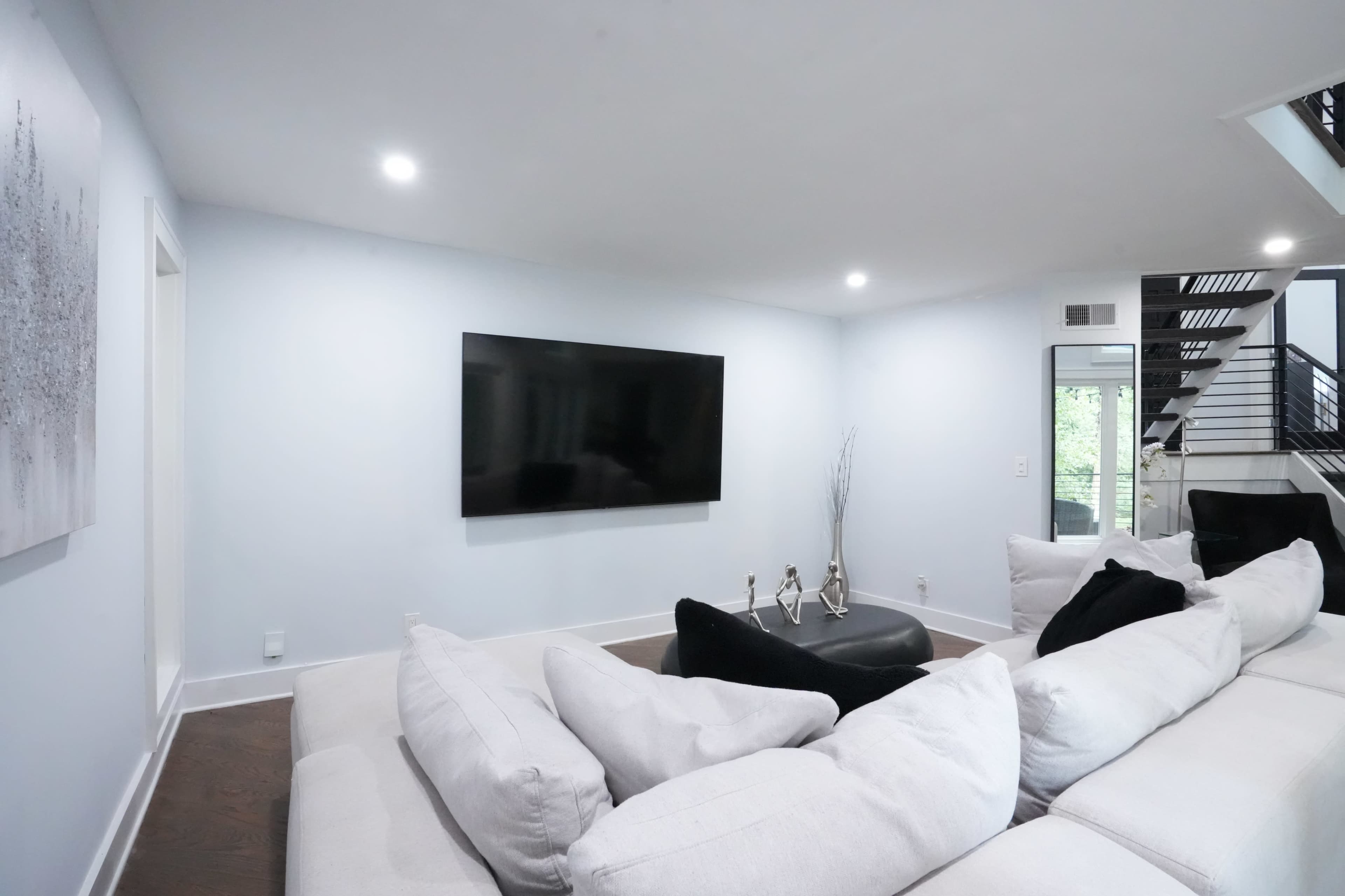 The image shows a modern living room with a large flat-screen television mounted on the wall, a light-colored sectional sofa, and a decorative mirror reflecting a staircase.