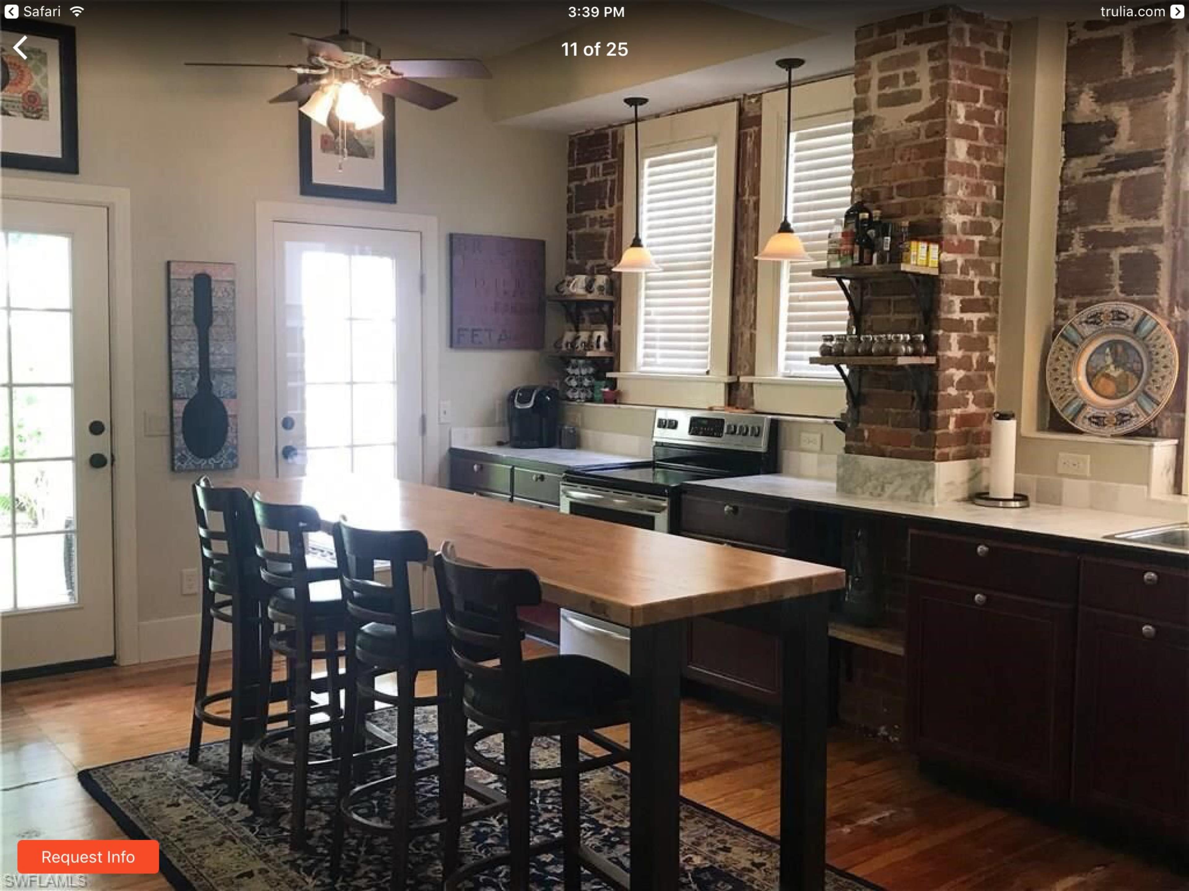 A kitchen with exposed brick walls, wooden cabinetry, a central island with four bar stools, and large windows letting in natural light.