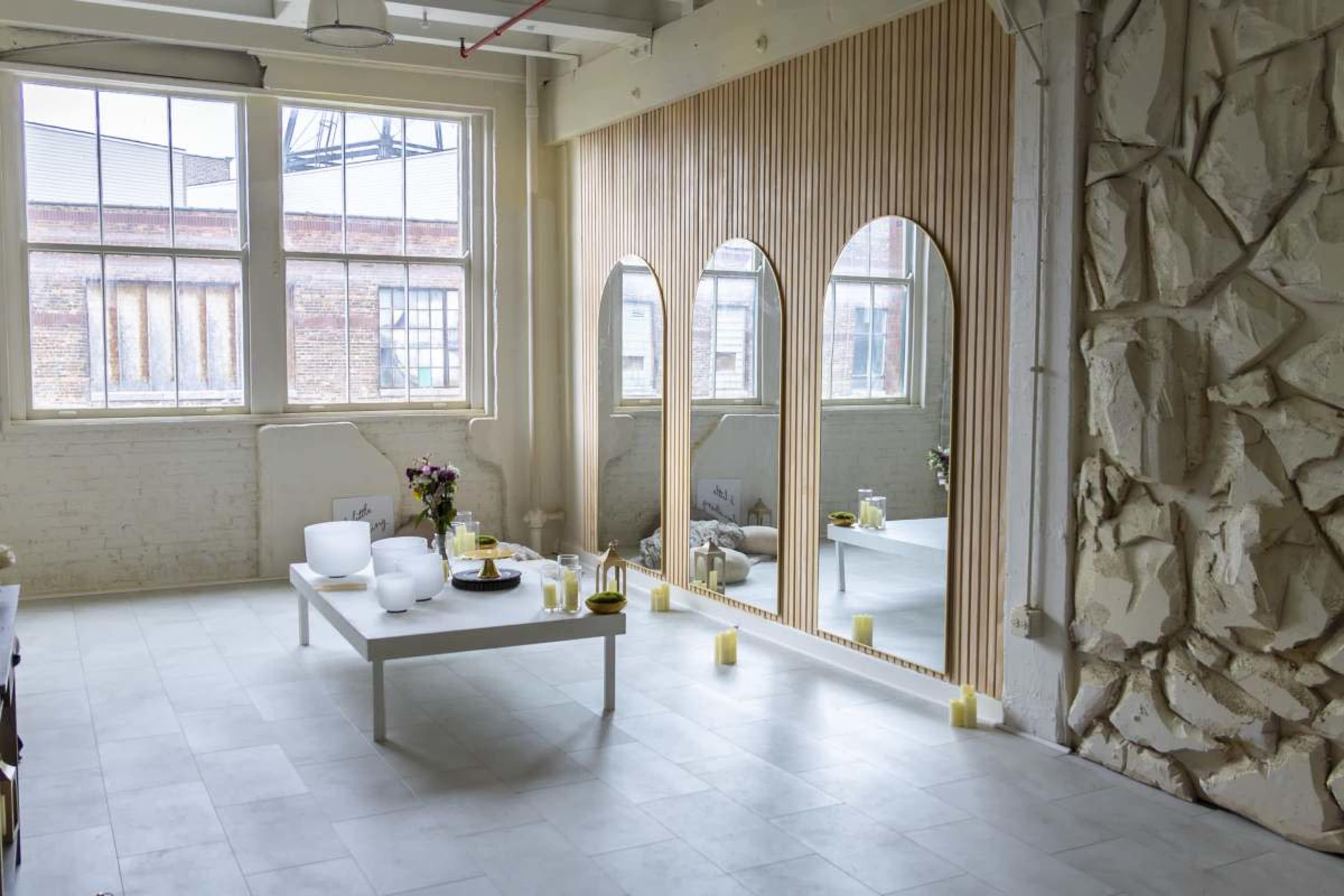The image shows a bright, airy room with large windows, a modern table set with various decorative items, and a unique mirror feature along one wall.