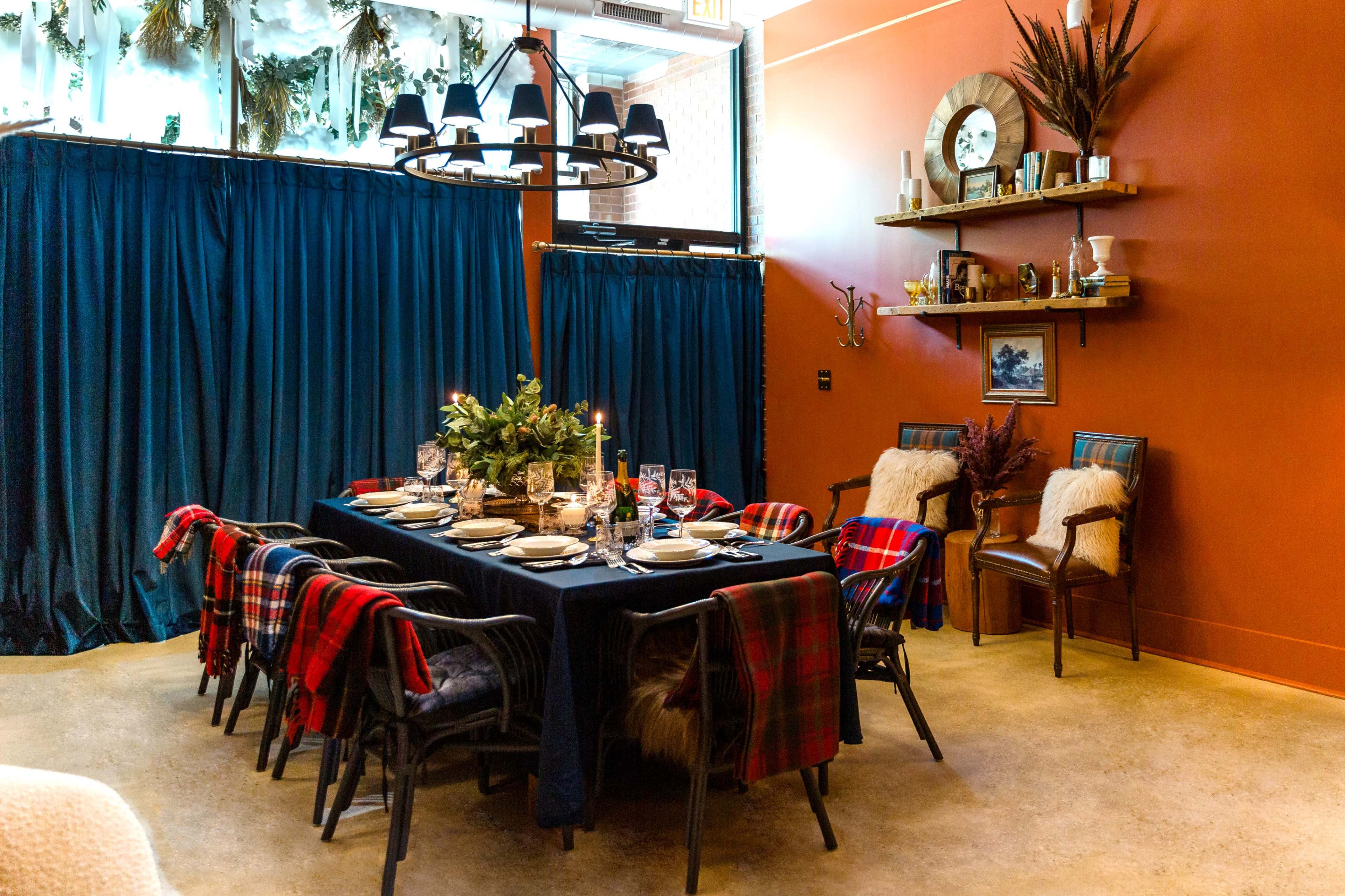 A long dining table is set with dishes and glasses, surrounded by chairs draped with plaid blankets, and a backdrop of a bright orange wall and dark curtains.