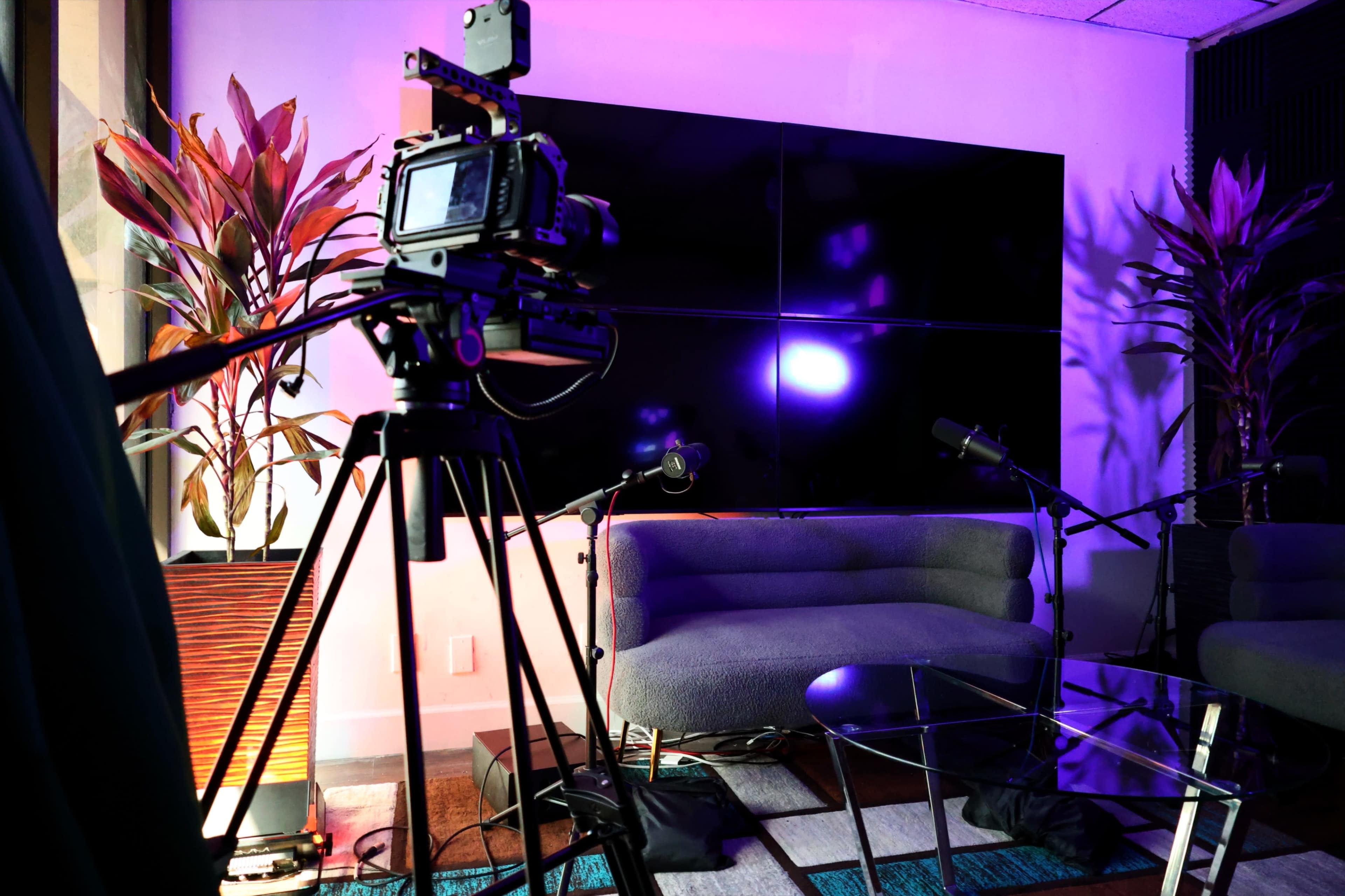 A camera on a tripod is positioned in front of a modern seating area with two microphones and a large screen in a dimly lit room.