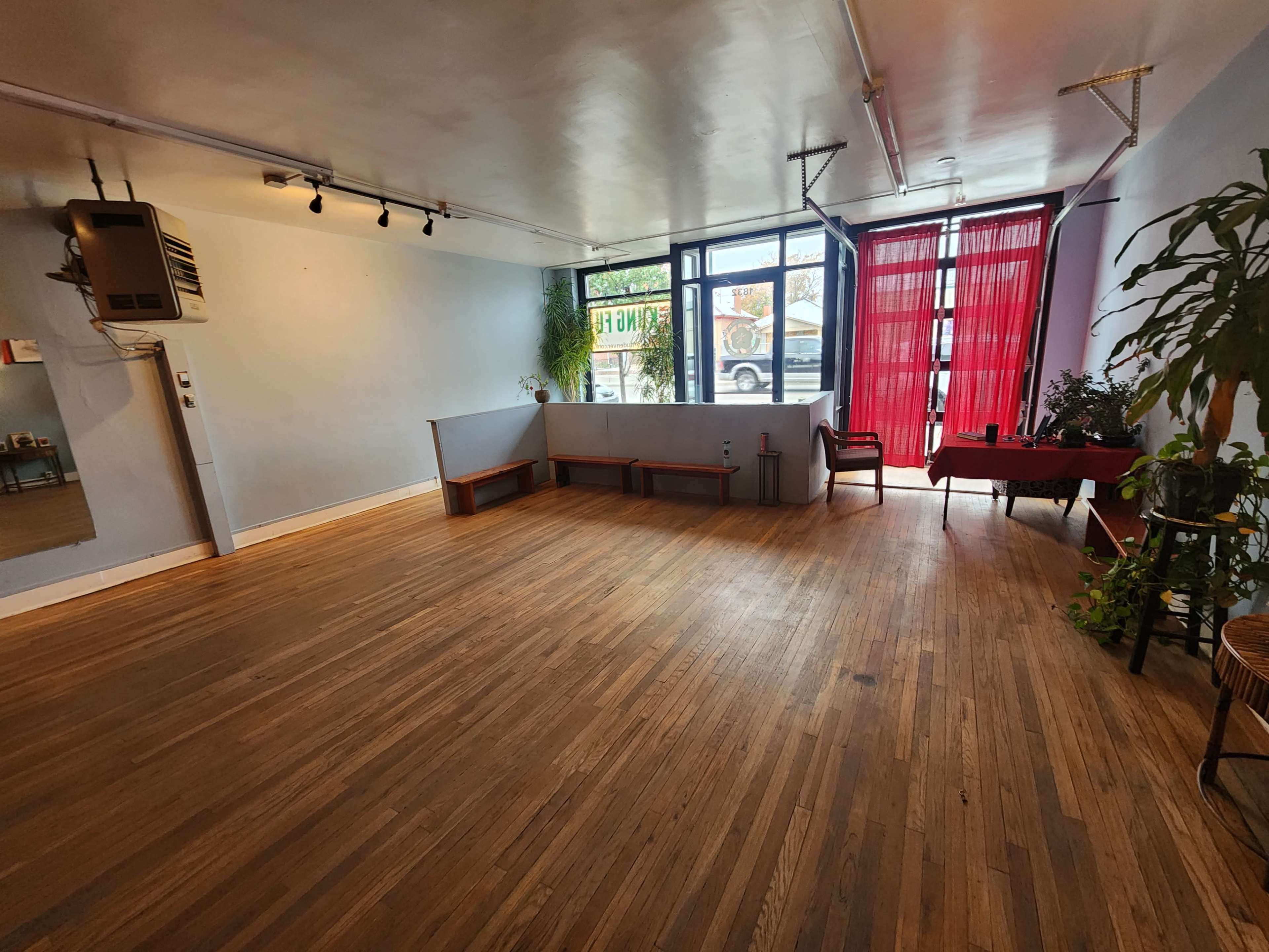 A spacious, well-lit room with wooden flooring, featuring a minimalist layout, large windows, and potted plants.