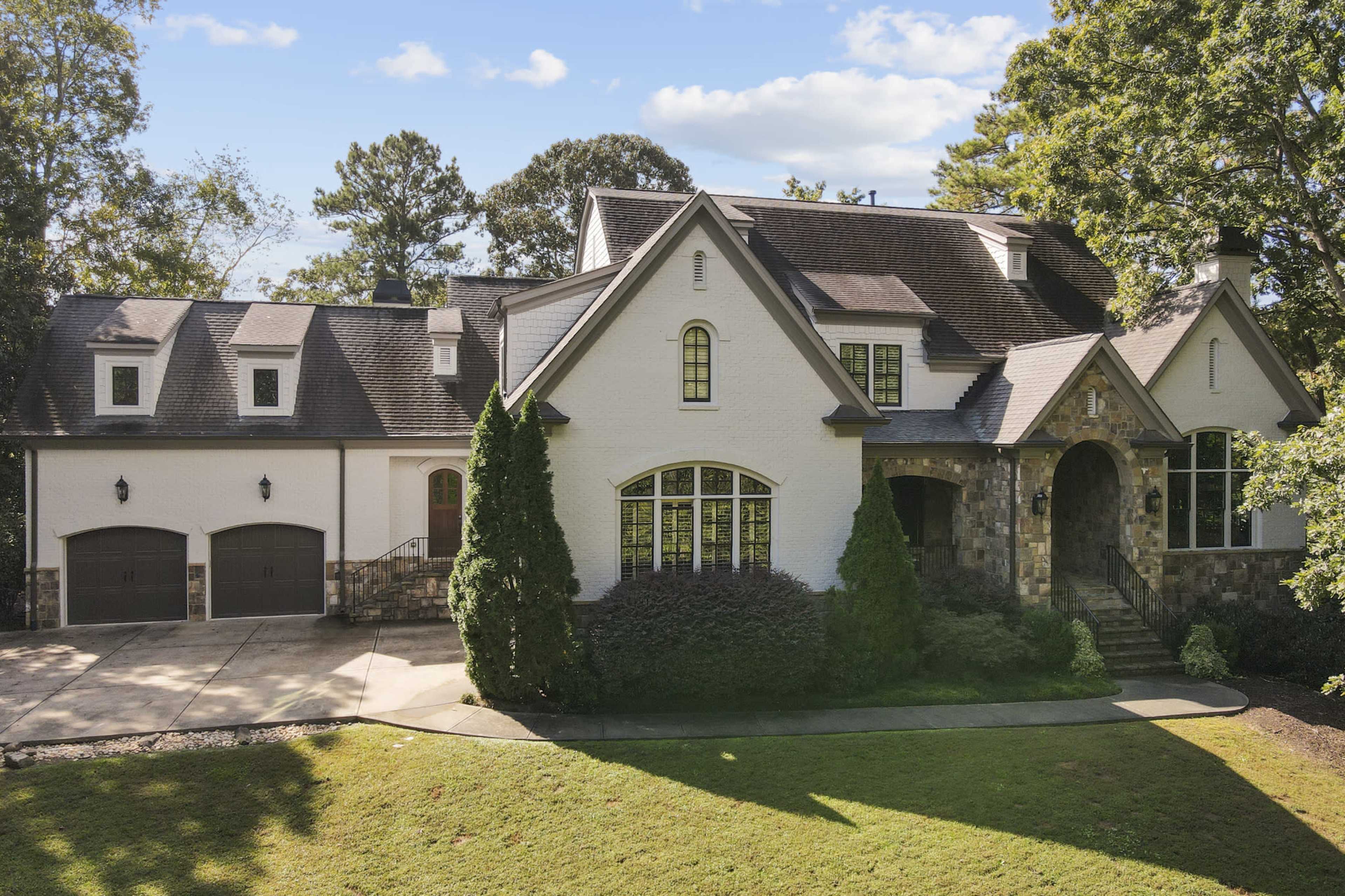 A large, modern house with a stone and stucco exterior, surrounded by greenery and featuring two garage doors.