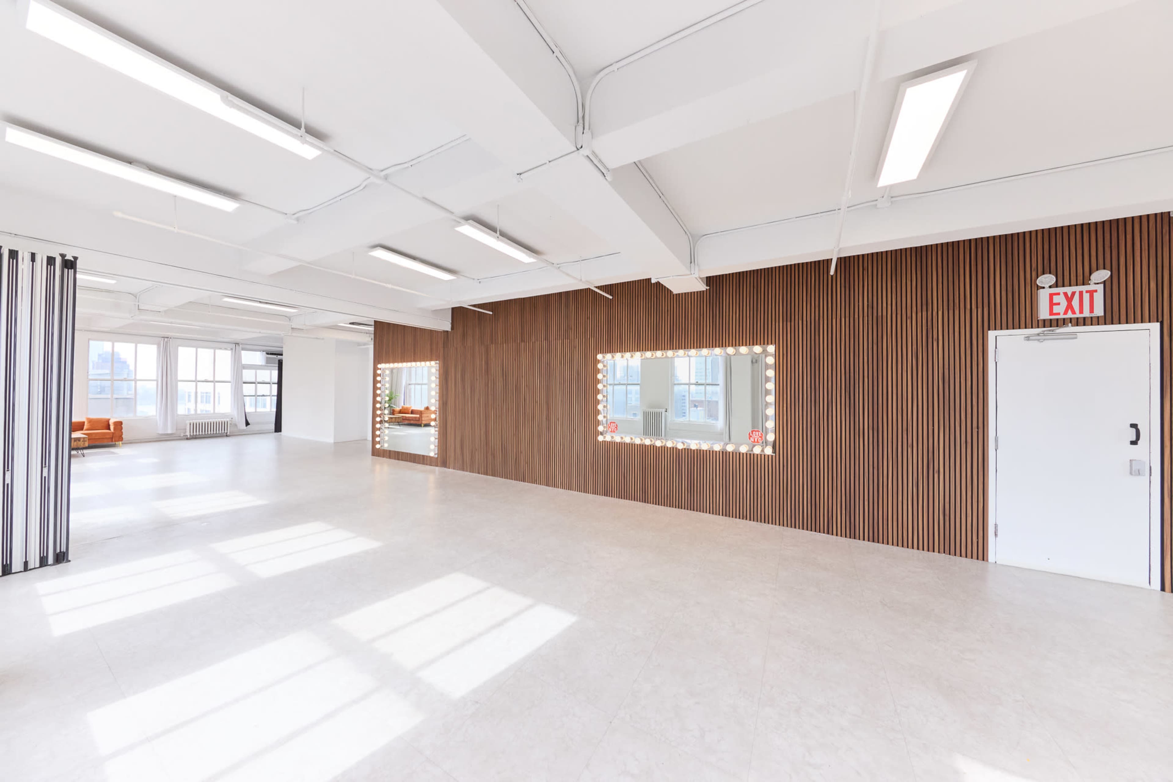 The image features a bright, spacious room with a wooden accent wall and a large mirror surrounded by lights.