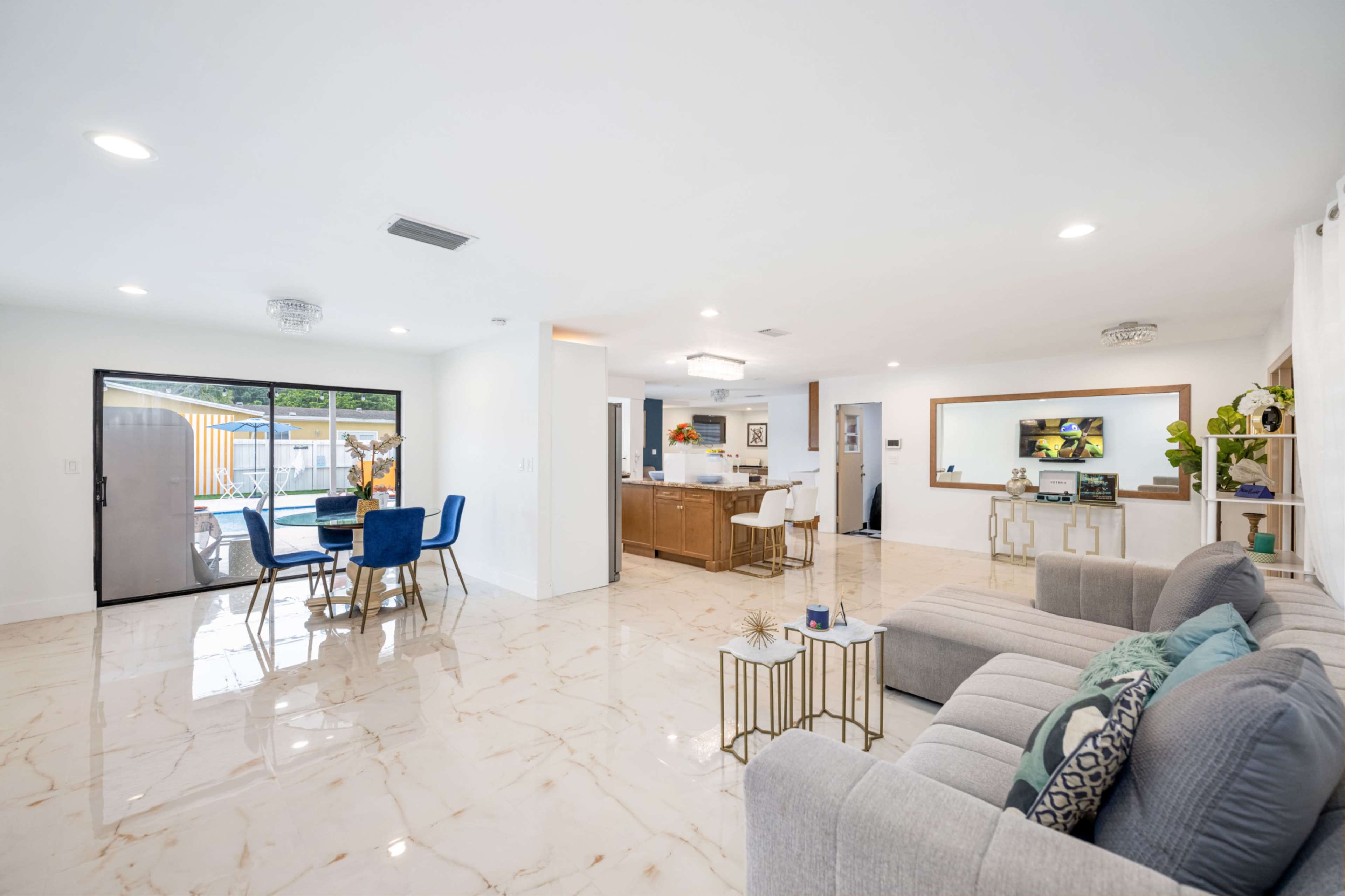 The image shows a modern living space with a combined dining and lounge area, featuring a large window, a kitchen in the background, and marble flooring.