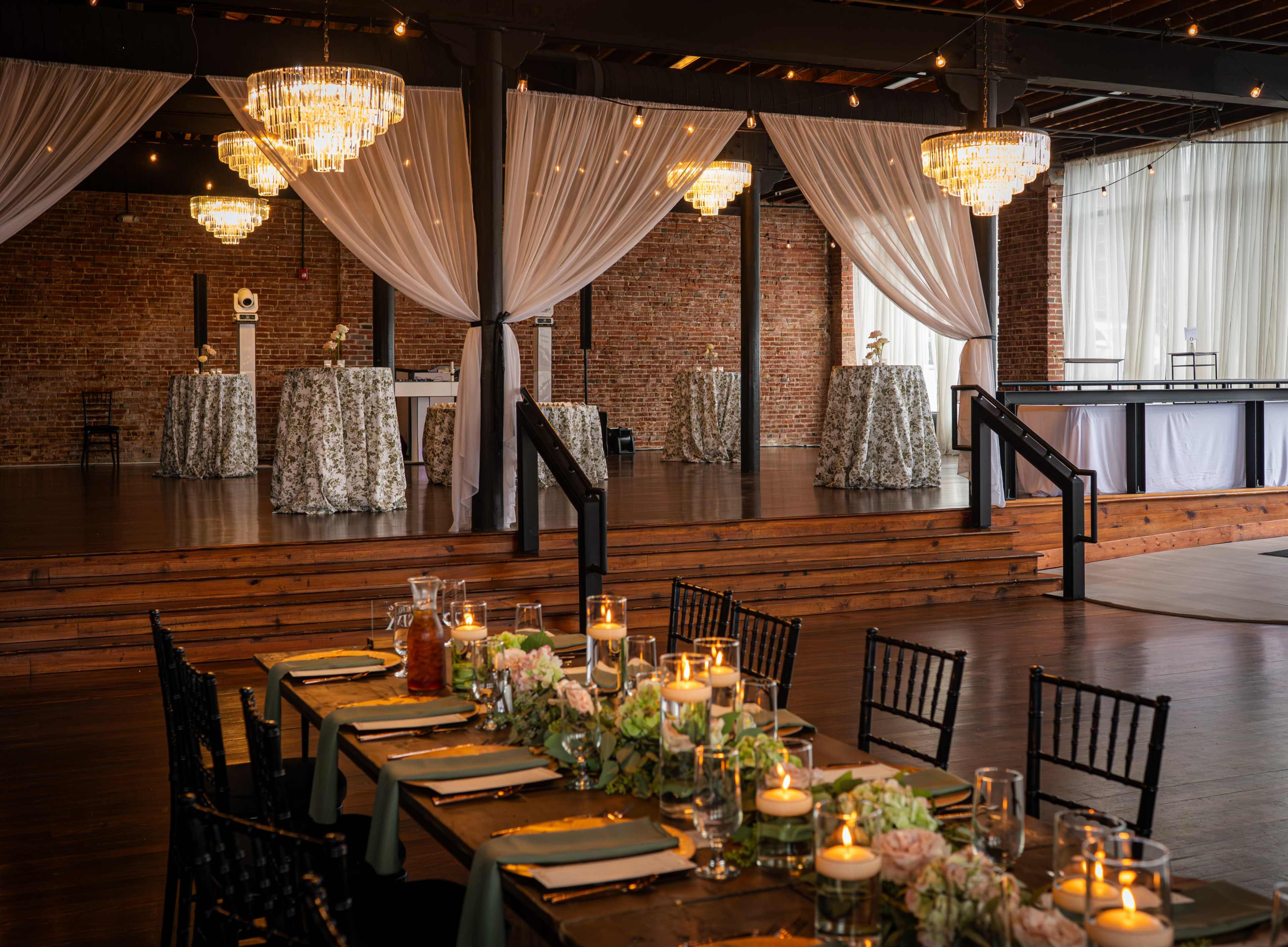 The image shows an elegant event space with wooden flooring, a stage area, and tables adorned with floral centerpieces and candles, all under draped fabric and chandeliers.
