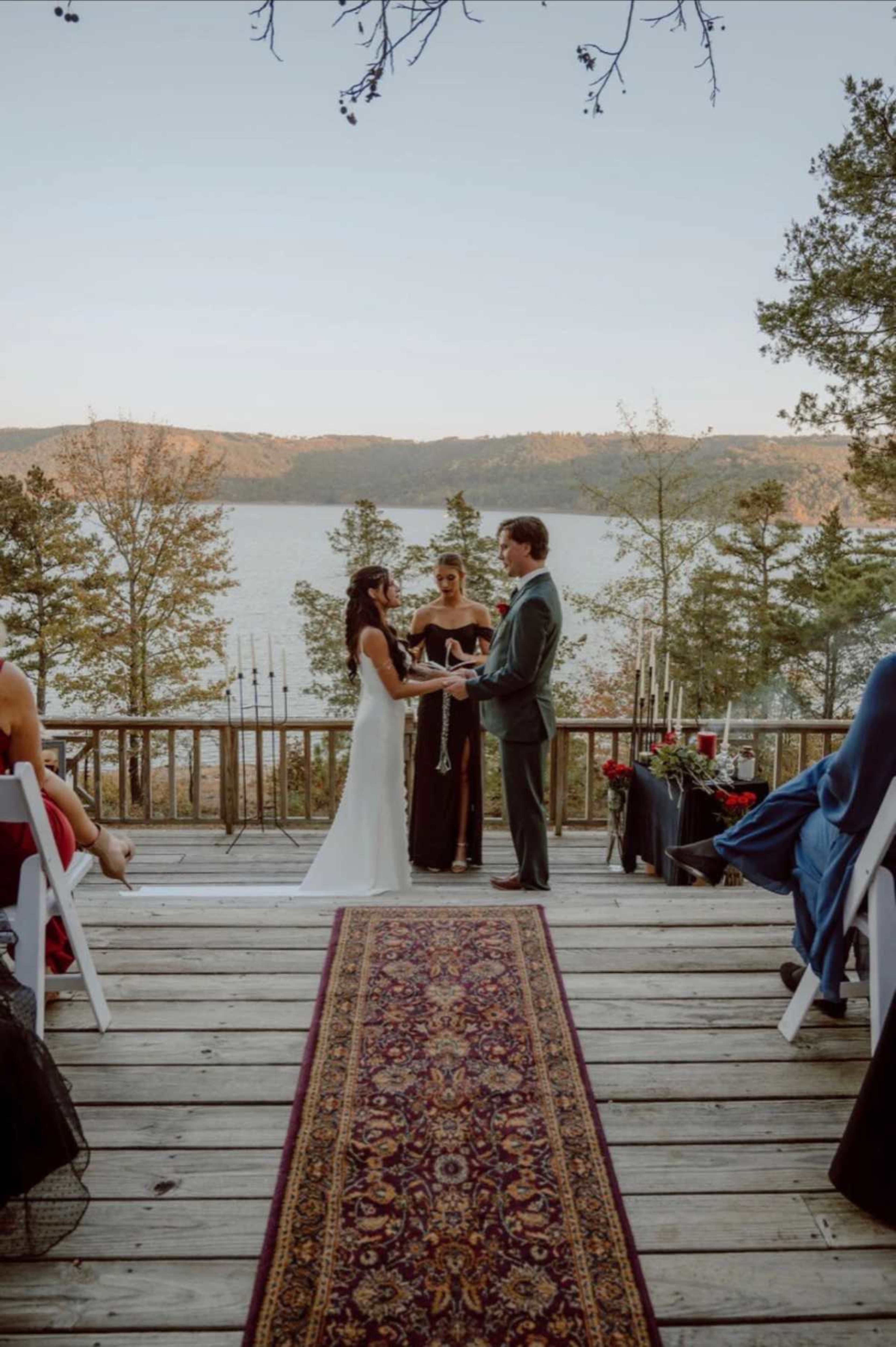 A couple stands on a wooden deck facing each other, holding hands during their wedding ceremony with a scenic lake and trees in the background.