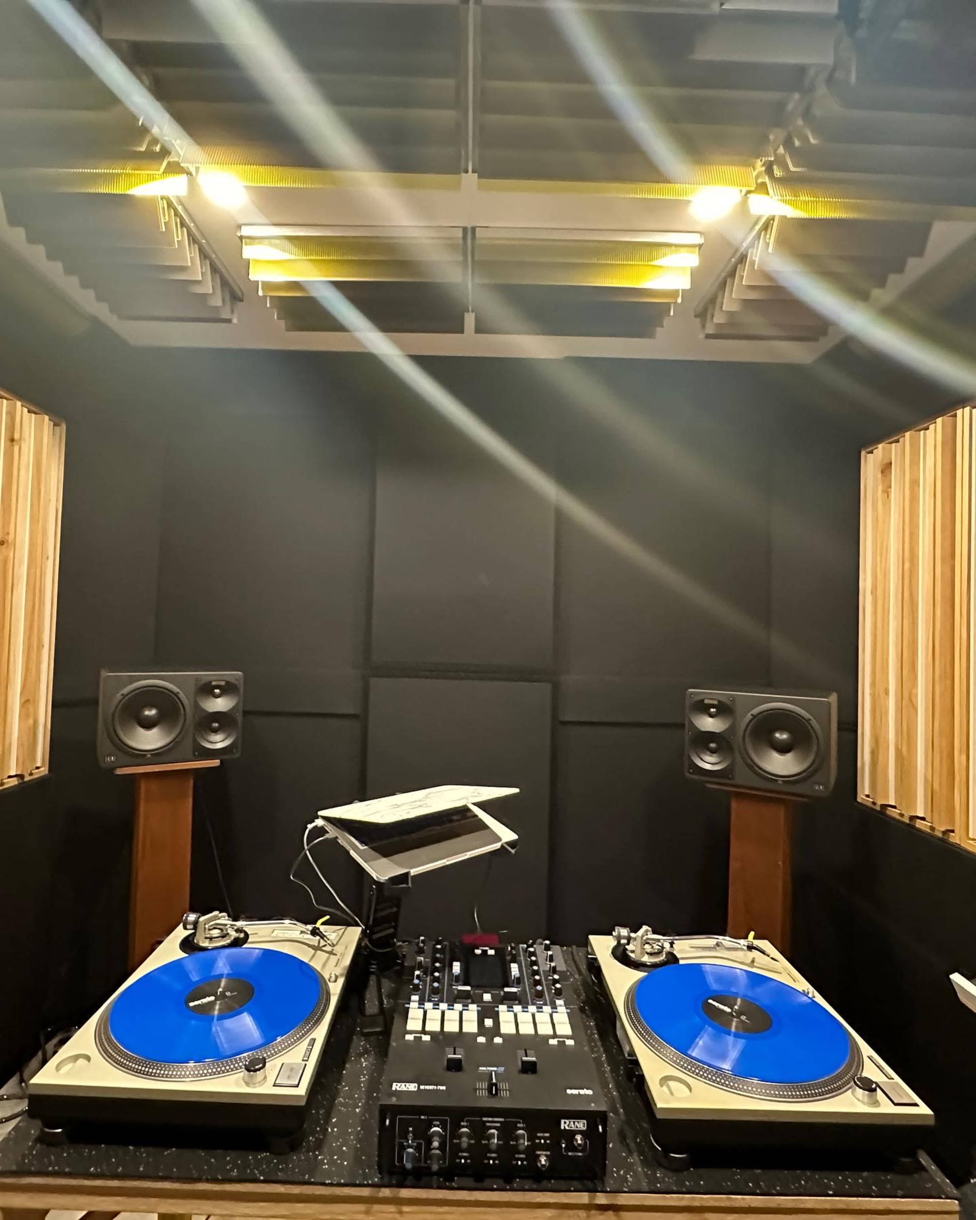 The image shows a DJ setup featuring two turntables with blue platters, a mixer in the center, and speakers mounted on either side within a soundproof studio booth.