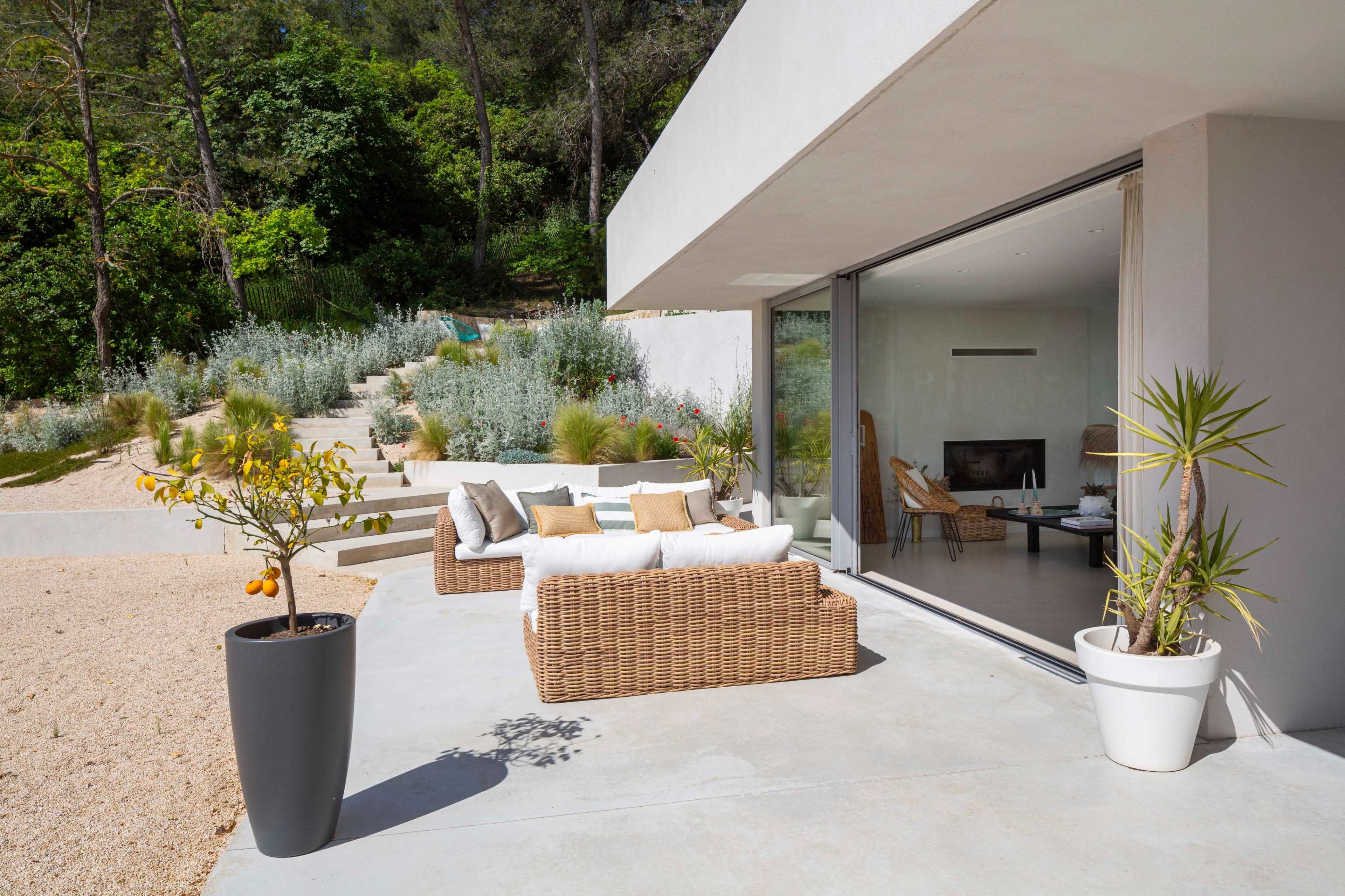 A modern outdoor seating area features a wicker sofa with cushions, surrounded by potted plants, overlooking a landscaped garden and pathway.