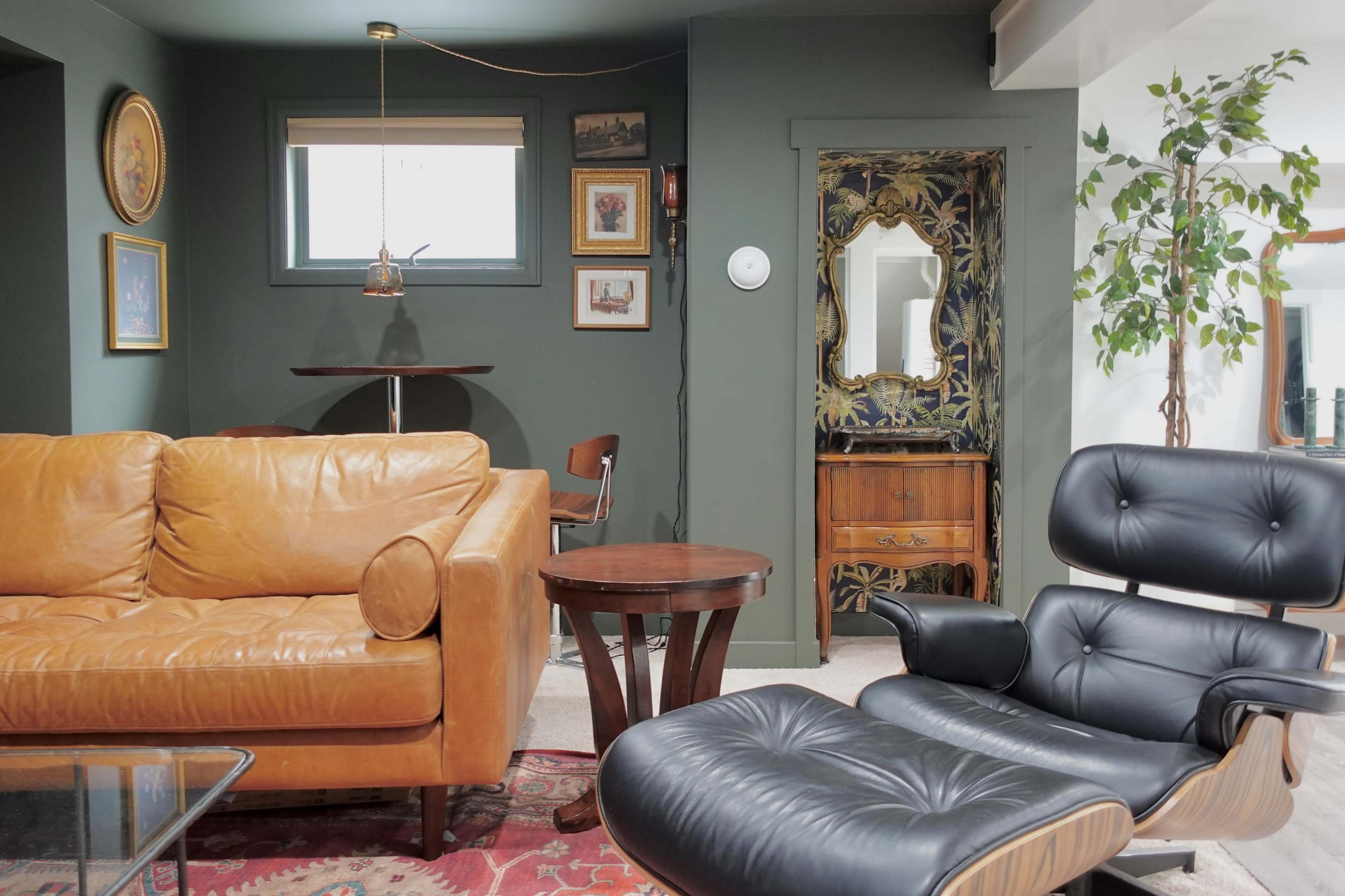 The image shows a cozy living room with a brown leather sofa, a round wooden table, a black lounge chair, and decorated walls featuring framed artwork and a tall indoor plant.