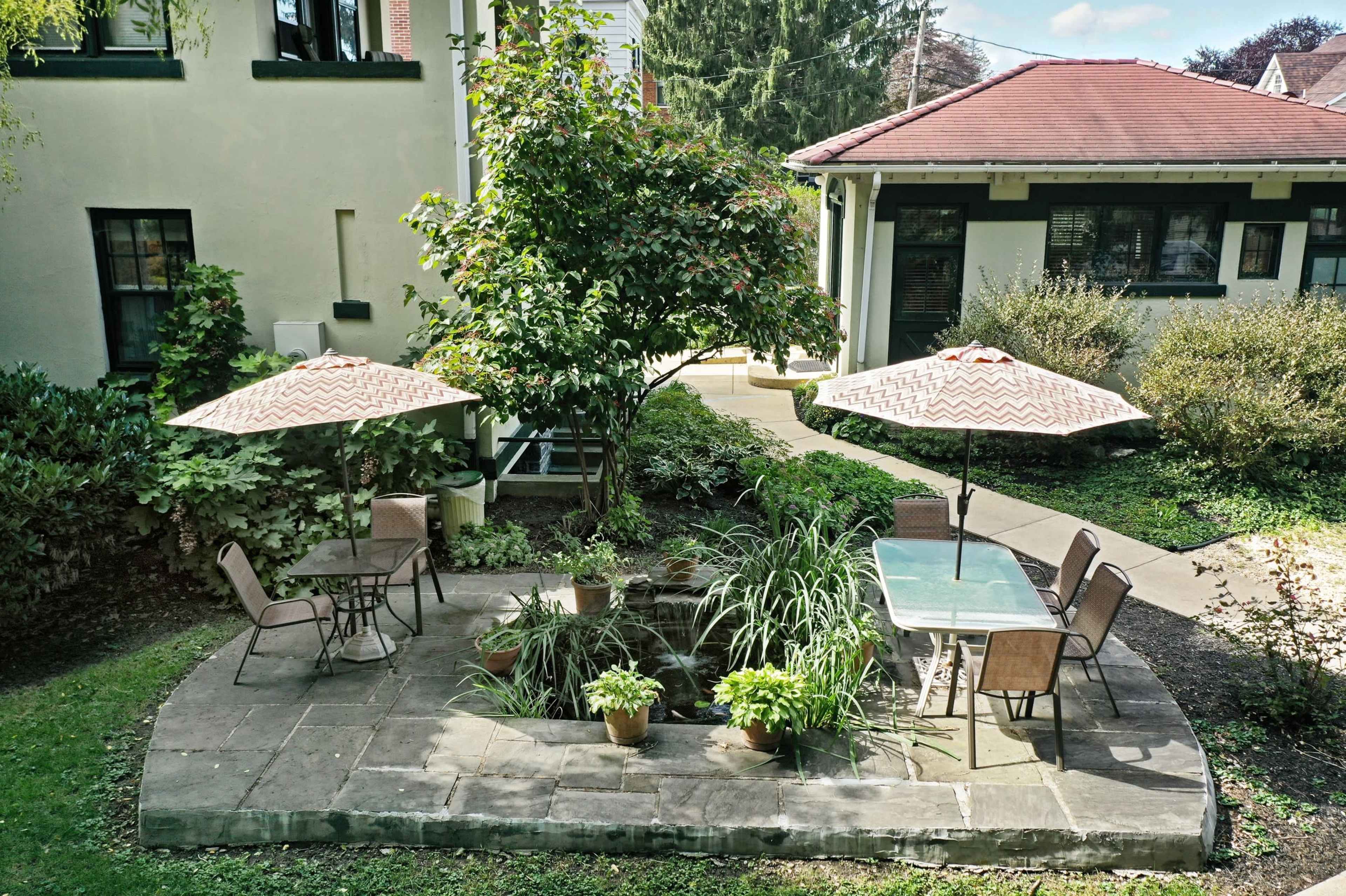 A patio featuring two shaded tables with chairs surrounded by greenery and a small pond.