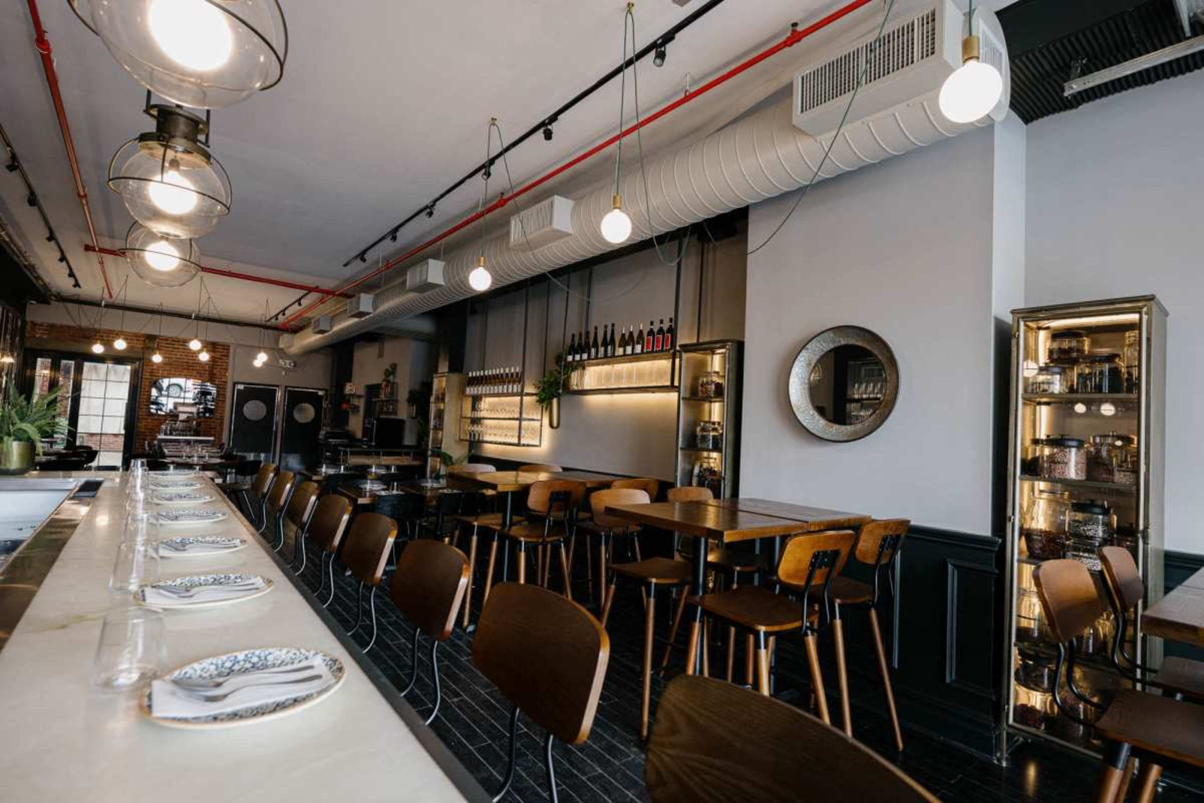 The image shows a modern restaurant interior featuring a long bar with round pendant lighting, wooden tables, and shelves displaying wine and other items along the walls.