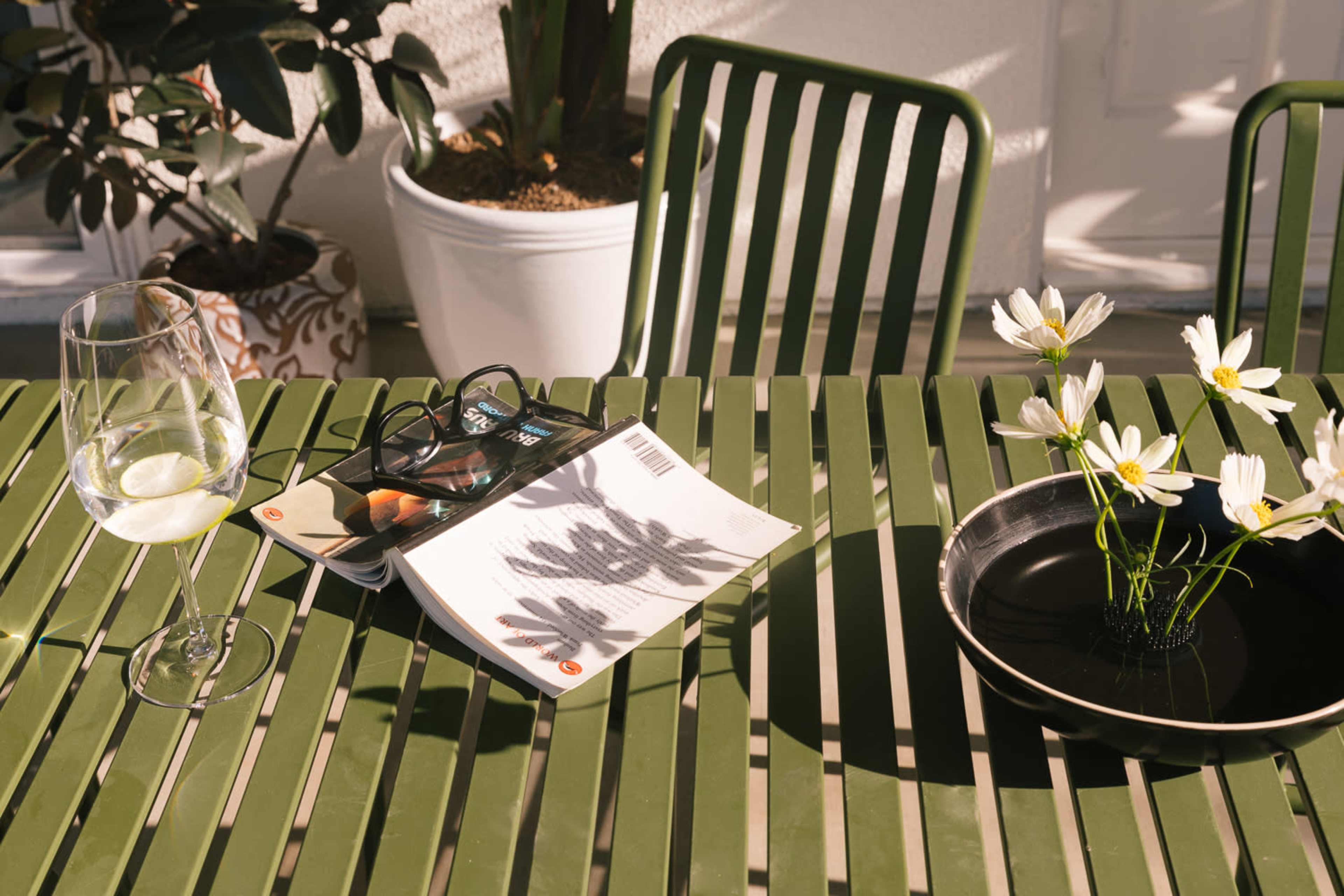 A glass of white wine and a pair of glasses lie on a magazine next to a bowl of flowers on a green table.
