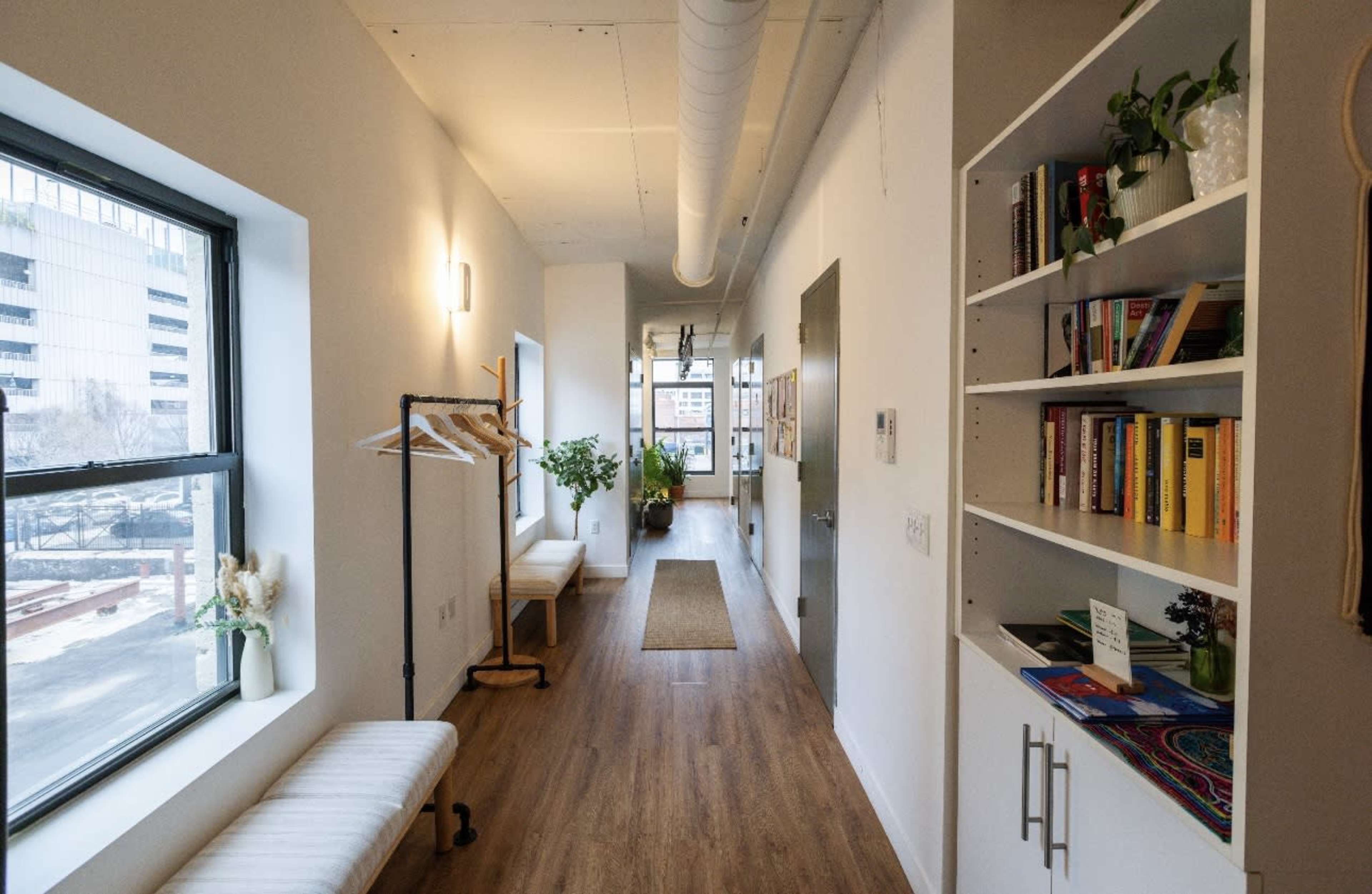 The image shows a well-lit corridor featuring a bench, plants, a bookshelf filled with books, and large windows along one side.