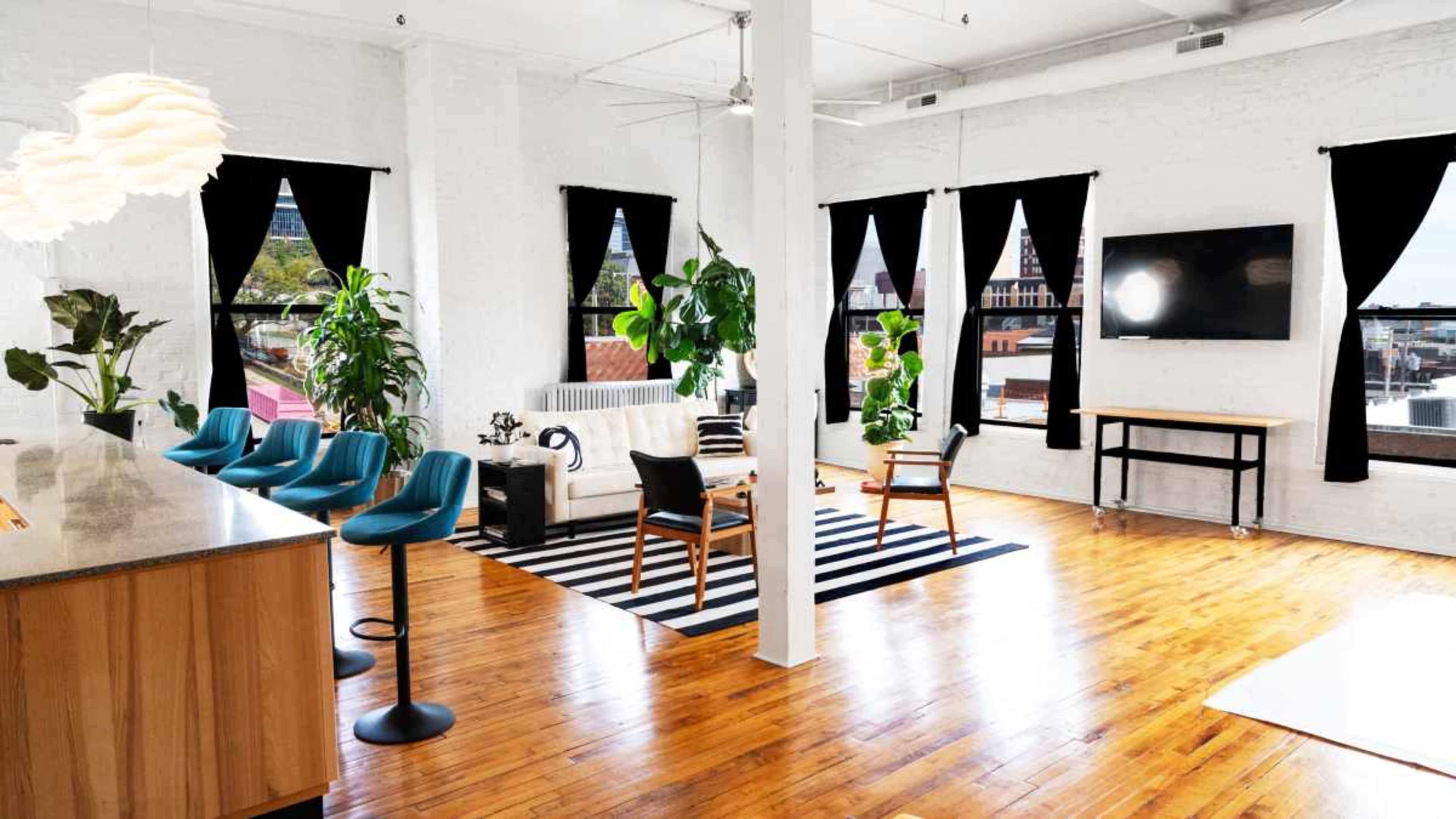The image shows a modern, open-plan living space featuring a kitchen area with bar stools, a sitting area with a sofa, and large windows dressed with black curtains, all highlighted by natural light streaming in.