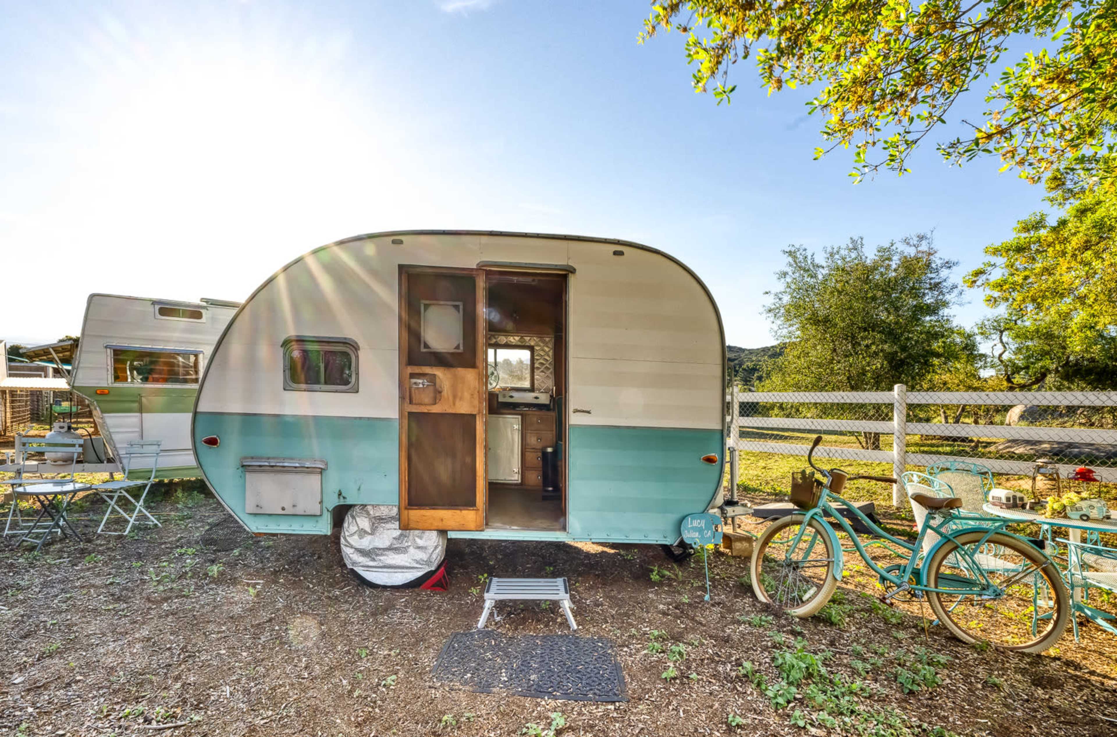 A vintage camper trailer is parked in a sunlit outdoor area, with green trees and a bicycle nearby.