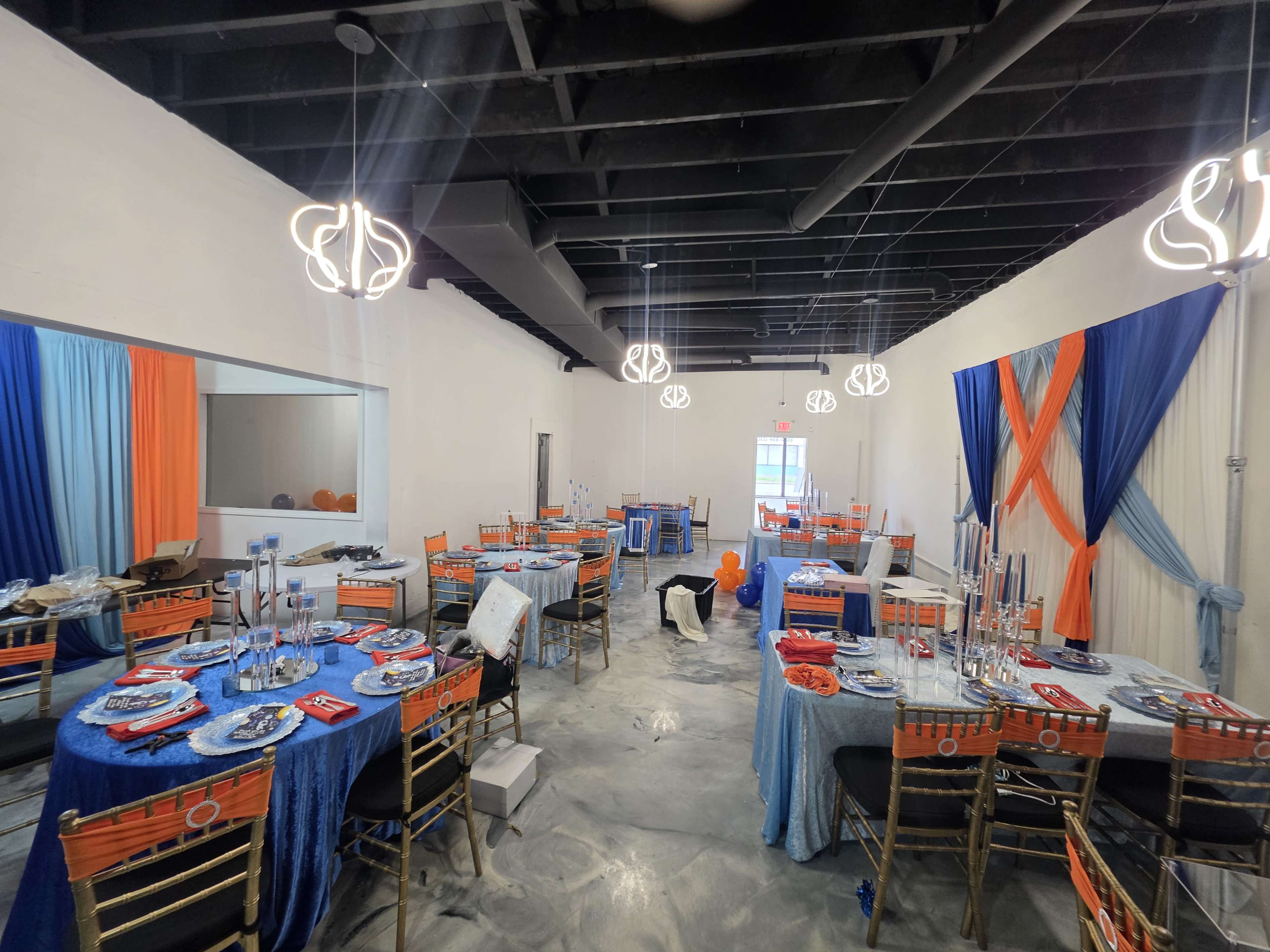 The image shows a large event space with multiple decorated tables, featuring blue and orange linens, candle holders, and hanging light fixtures.