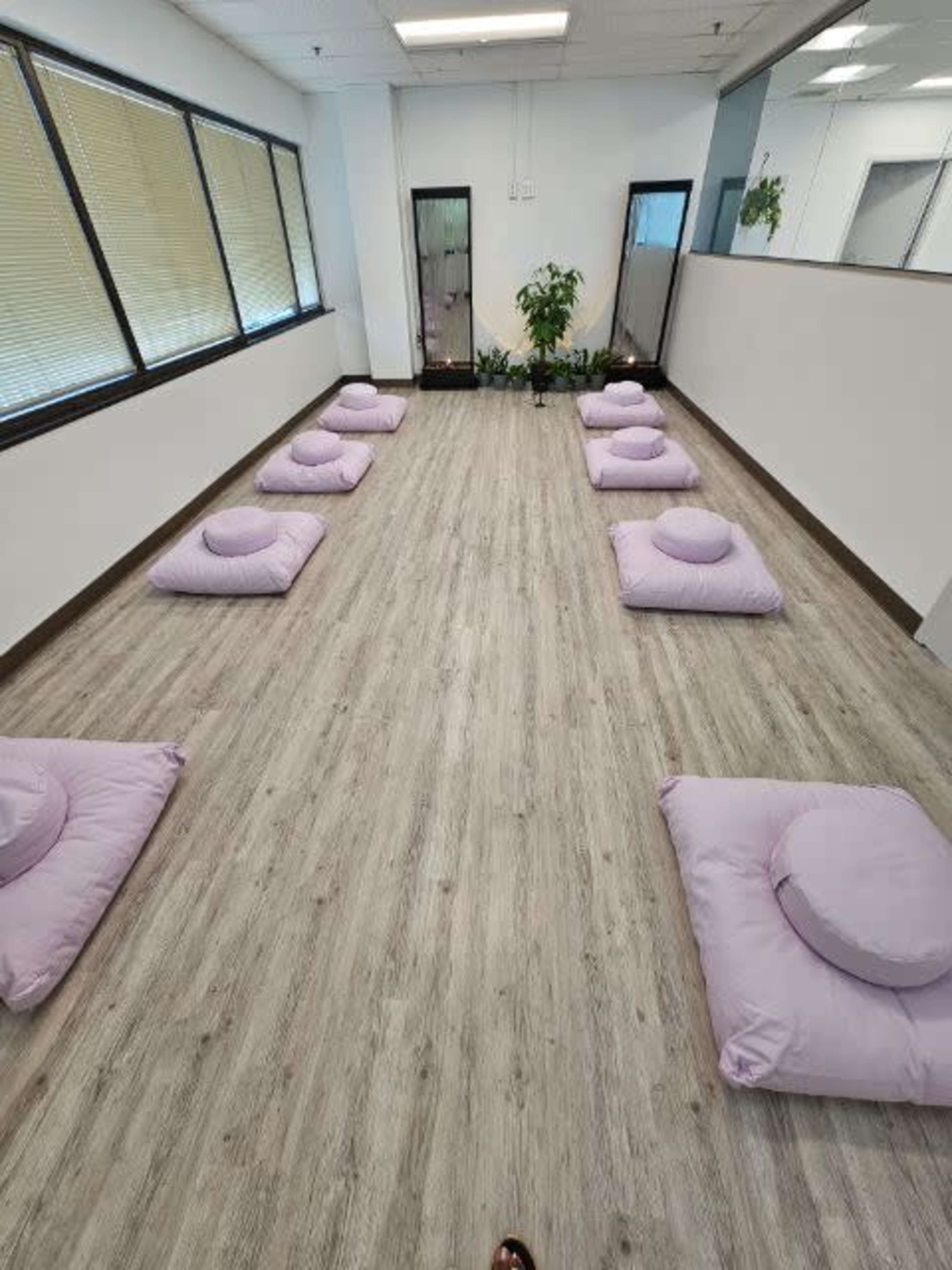 A spacious, minimalist room with several lavender cushions arranged on a wooden floor, flanked by plants and large windows.