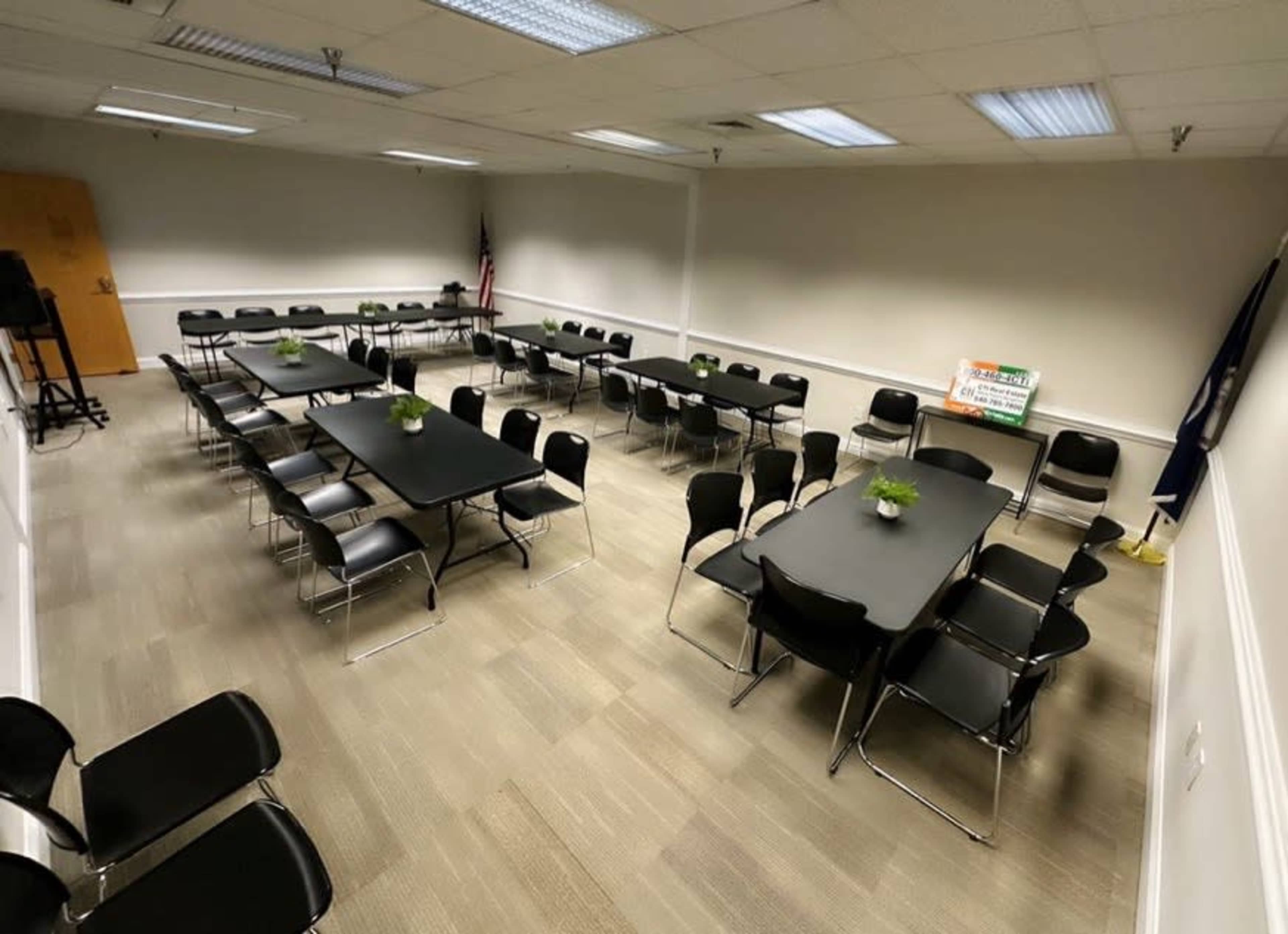 A spacious meeting room features several long tables arranged in a symmetrical layout with chairs surrounding them, and small plants placed as centerpieces.