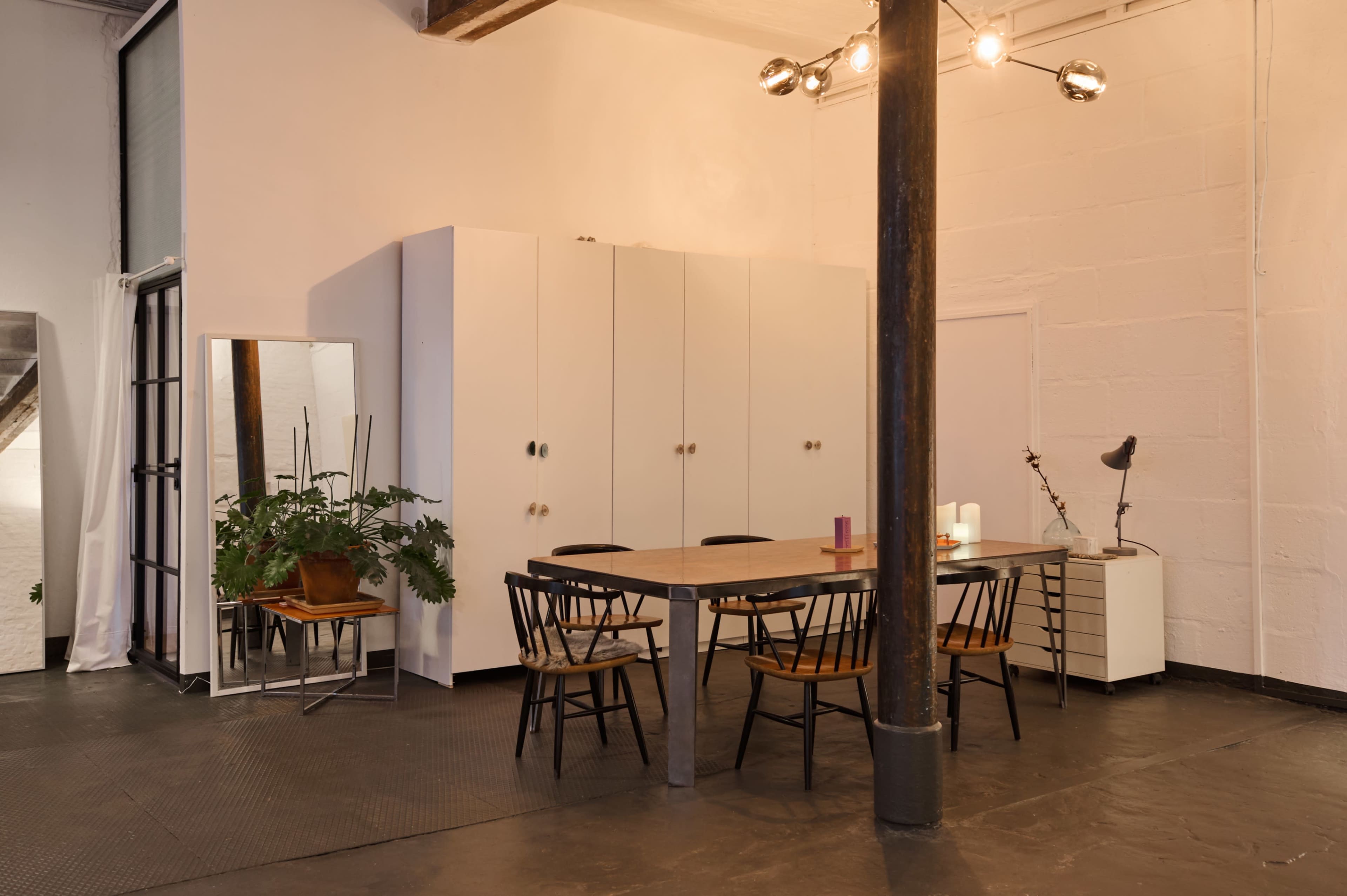 The image shows a minimalist dining area with a wooden table surrounded by black chairs, a large mirror, a potted plant, and storage cabinets against a white wall.