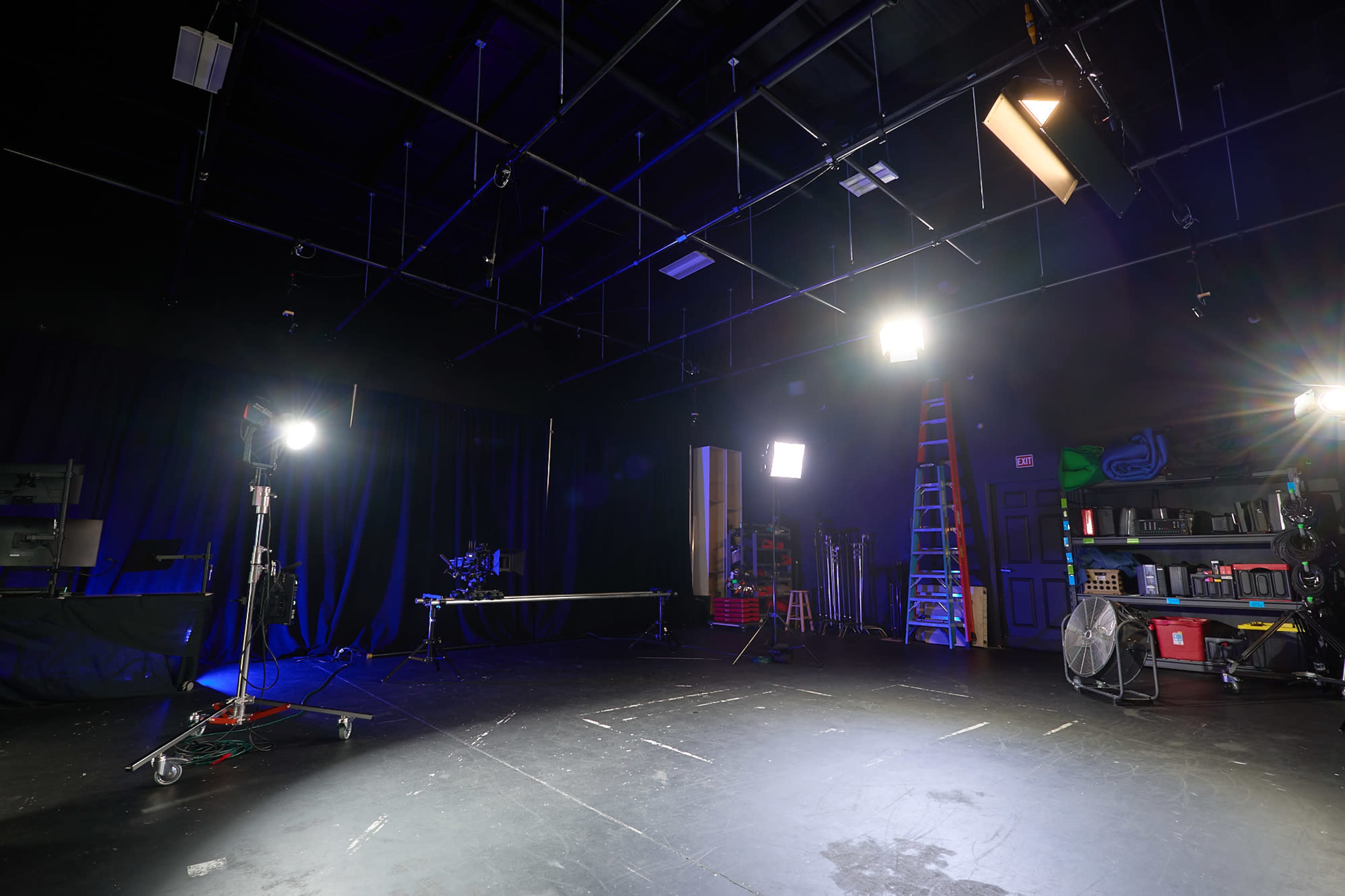 The image shows a dimly lit film studio with various lighting equipment, a tripod, and a ladder against the wall.