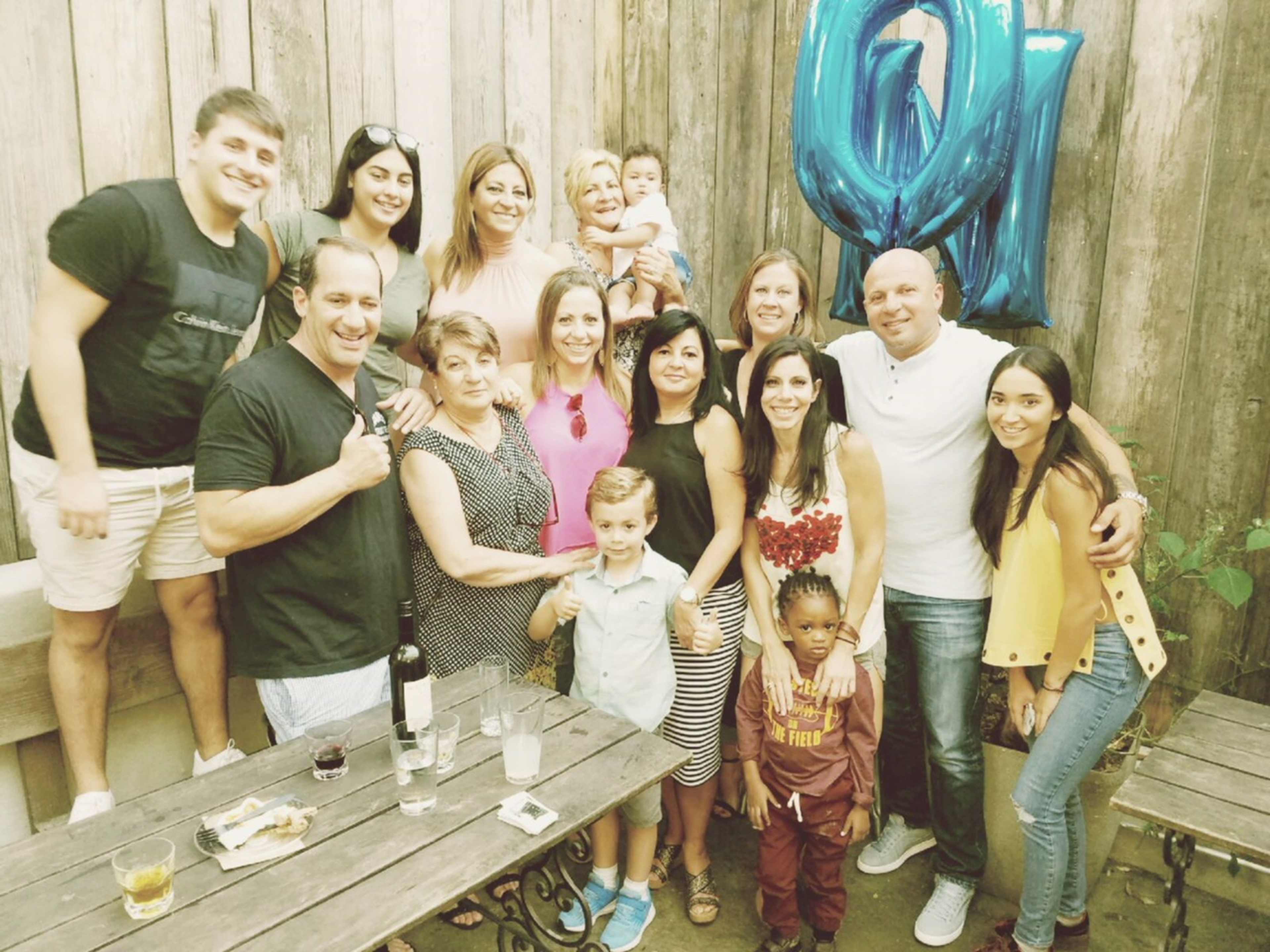 A group of people celebrates an occasion in a rustic outdoor setting with wooden walls and balloons.