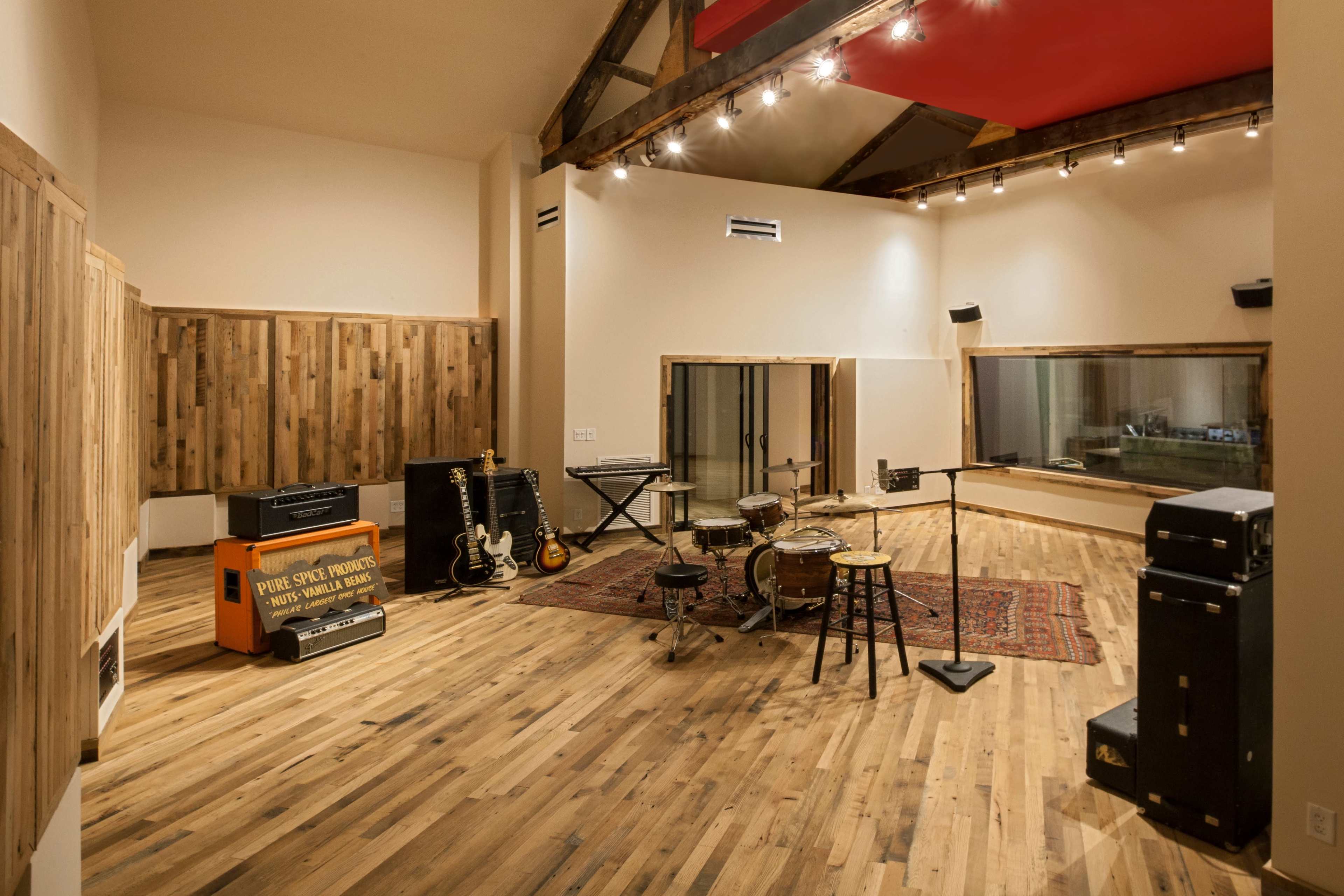 The image shows a well-lit music studio featuring various instruments, including guitars, a drum set, and sound equipment, arranged in a spacious wooden interior.