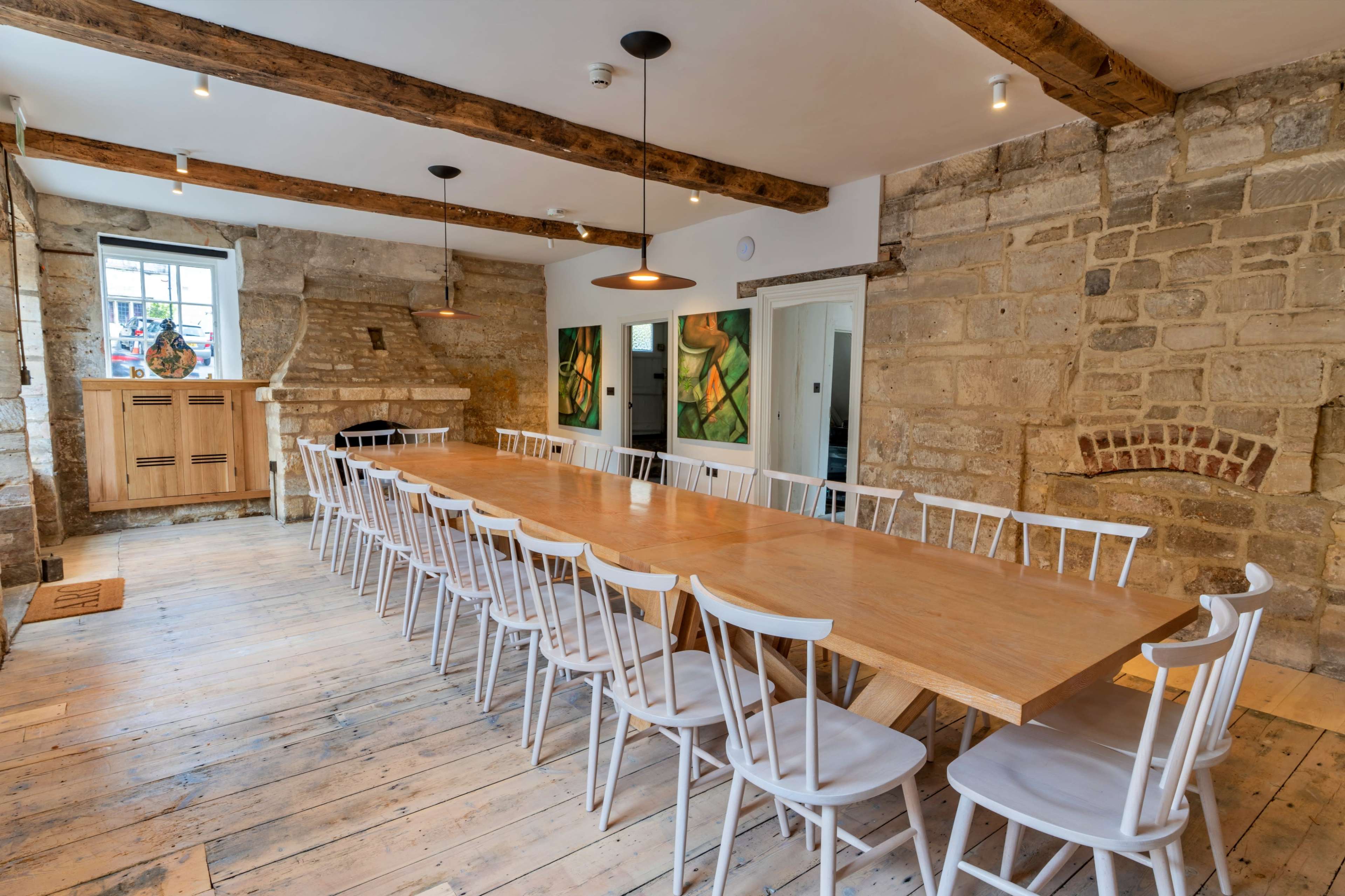 A large wooden table is set in a spacious room with exposed stone walls and wooden beam ceilings, surrounded by white chairs.