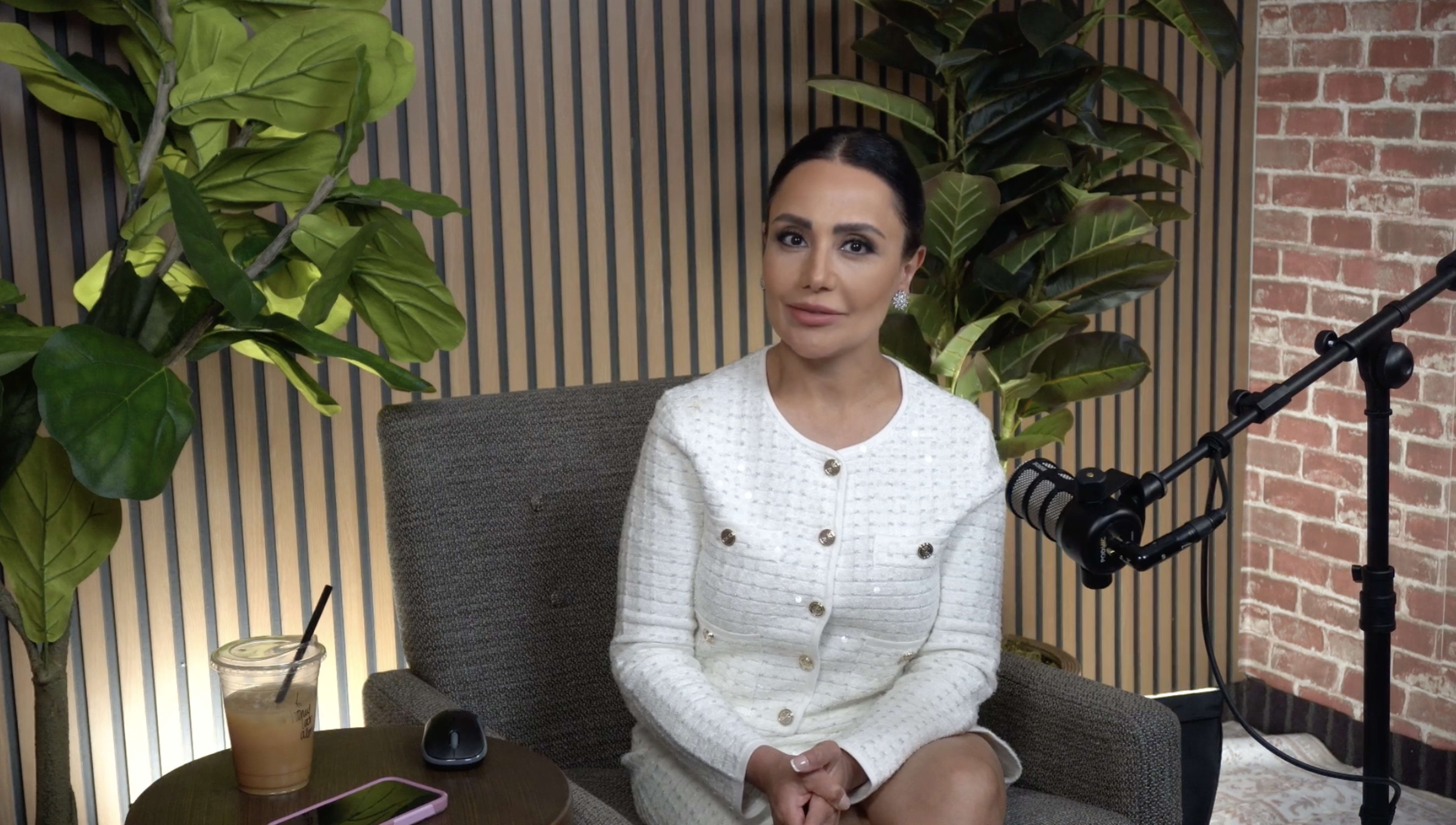 A woman sits on a chair in a room decorated with potted plants, facing the camera while wearing a white jacket with buttons.