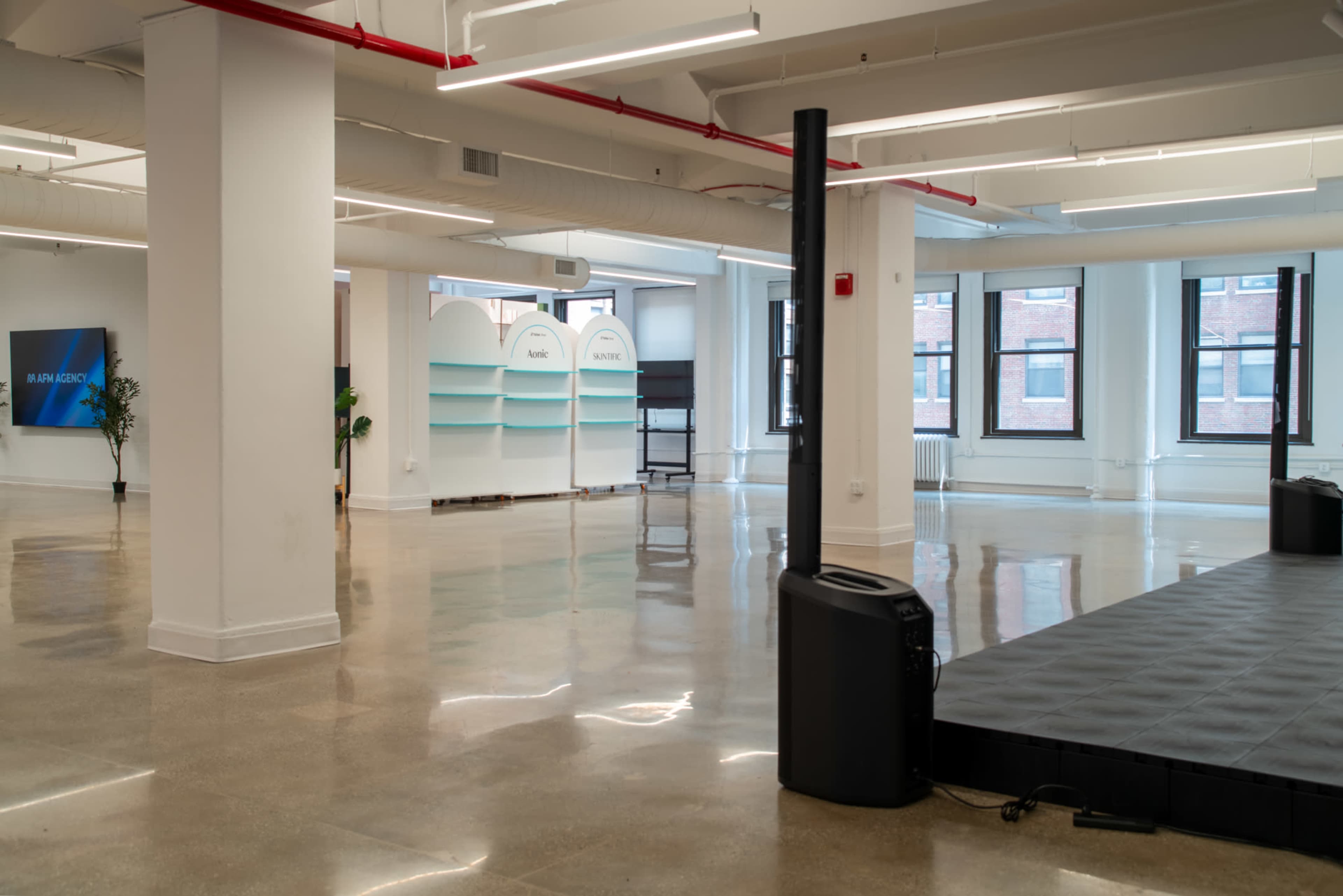 The image shows a spacious, empty room with polished concrete floors, featuring a small stage area and light-colored shelving units along the walls.