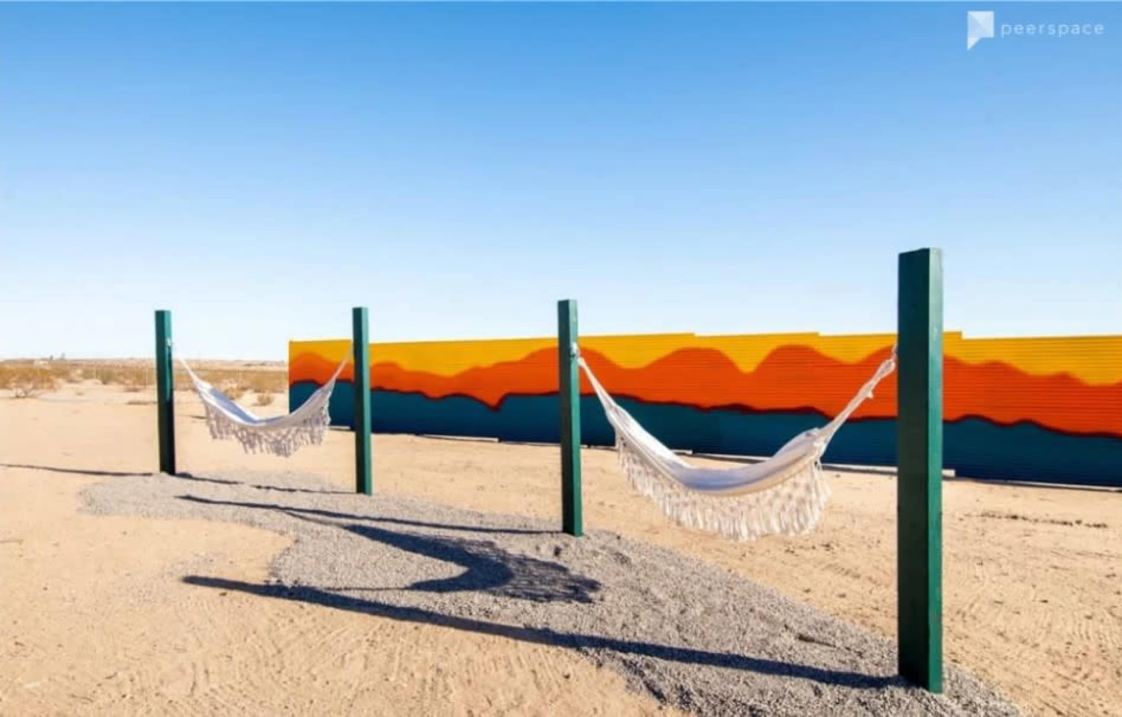 Two hammocks are suspended between green posts in a desert setting, with a colorful metal wall in the background.