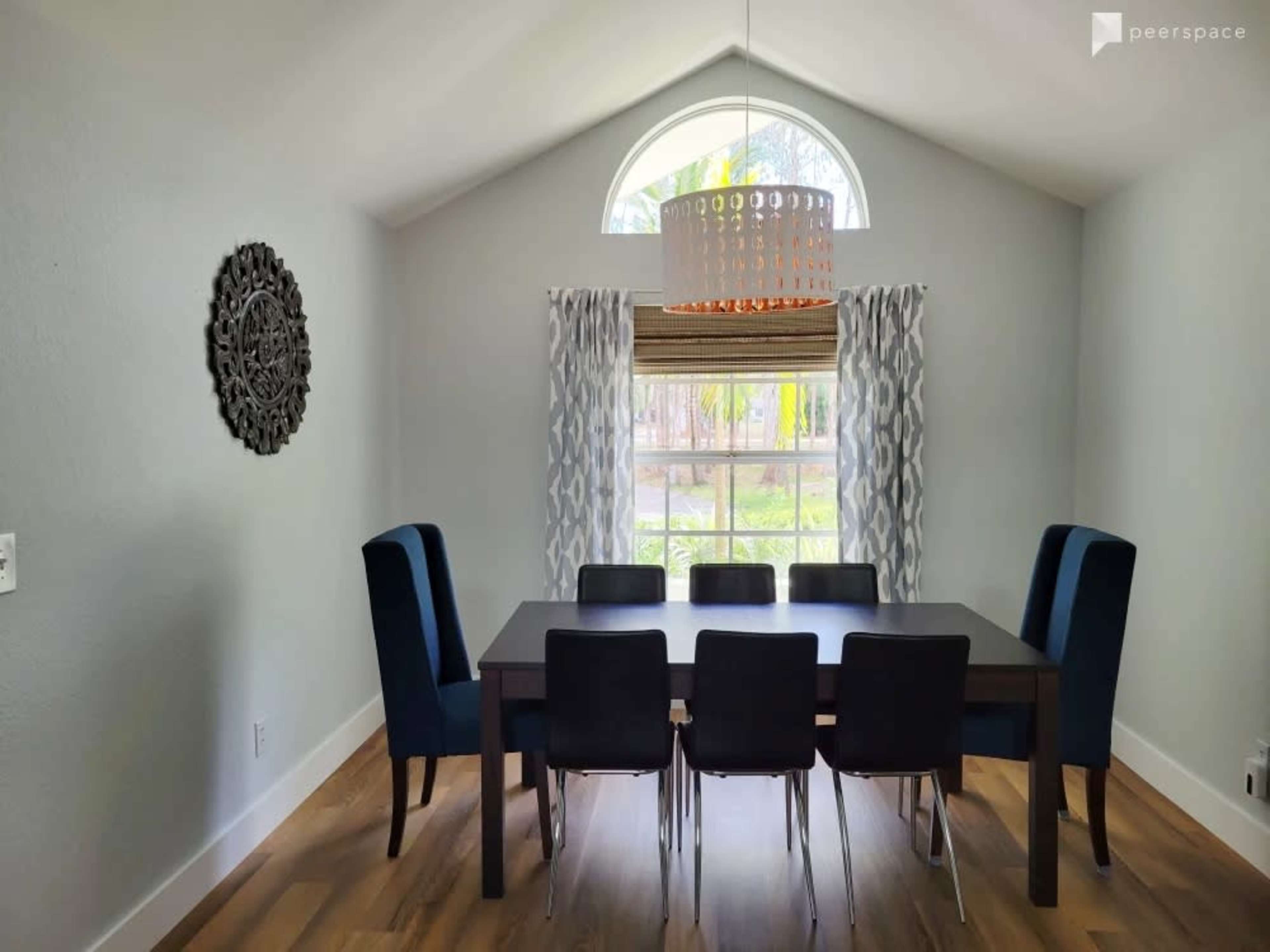 A modern dining room features a rectangular table surrounded by black chairs and blue upholstered armchairs, with a large window and decorative lighting above.