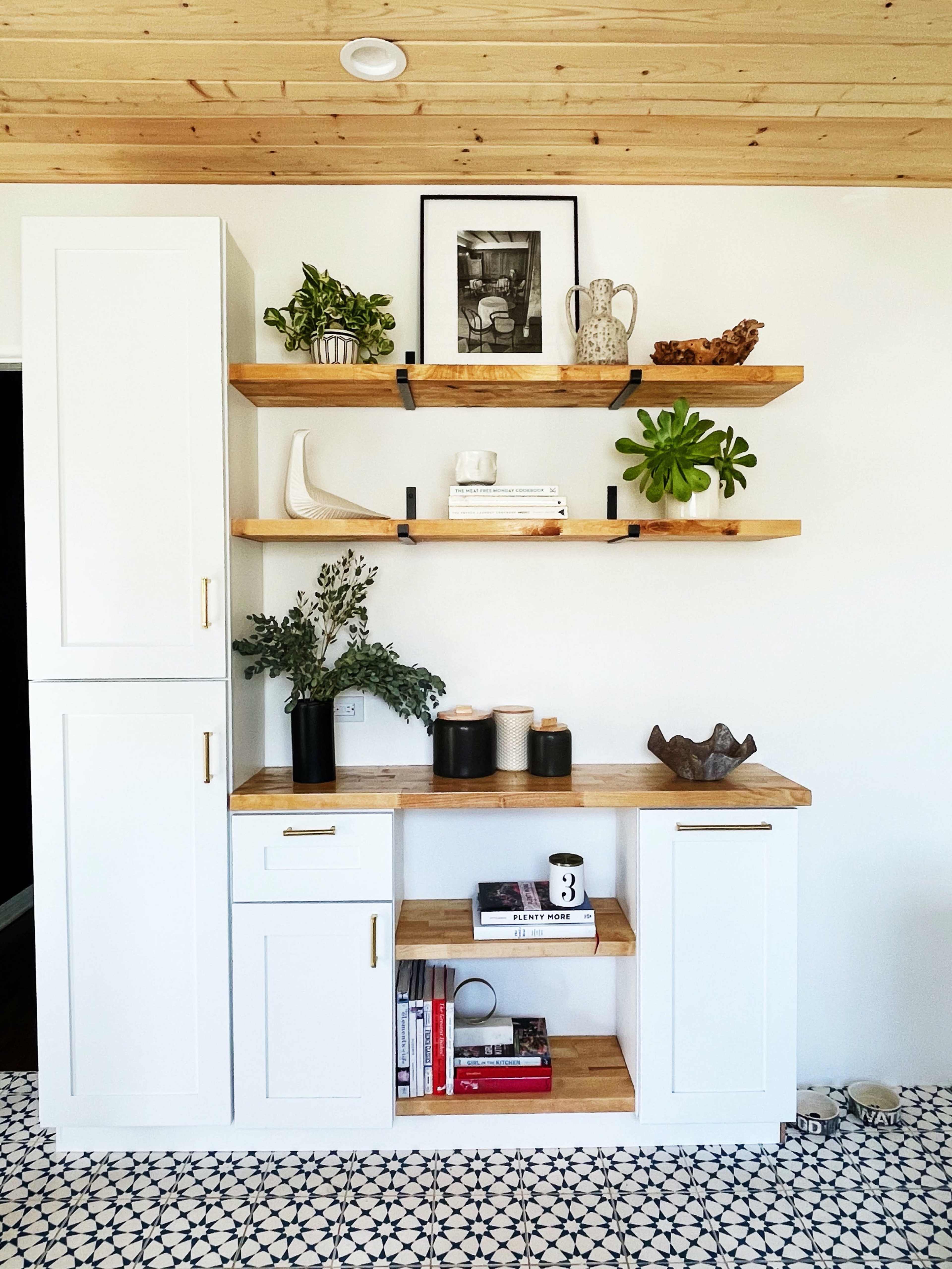 A minimalist interior featuring a white cabinetry unit with open wooden shelves, which hold various plants and decorative items, set against a patterned tile floor.