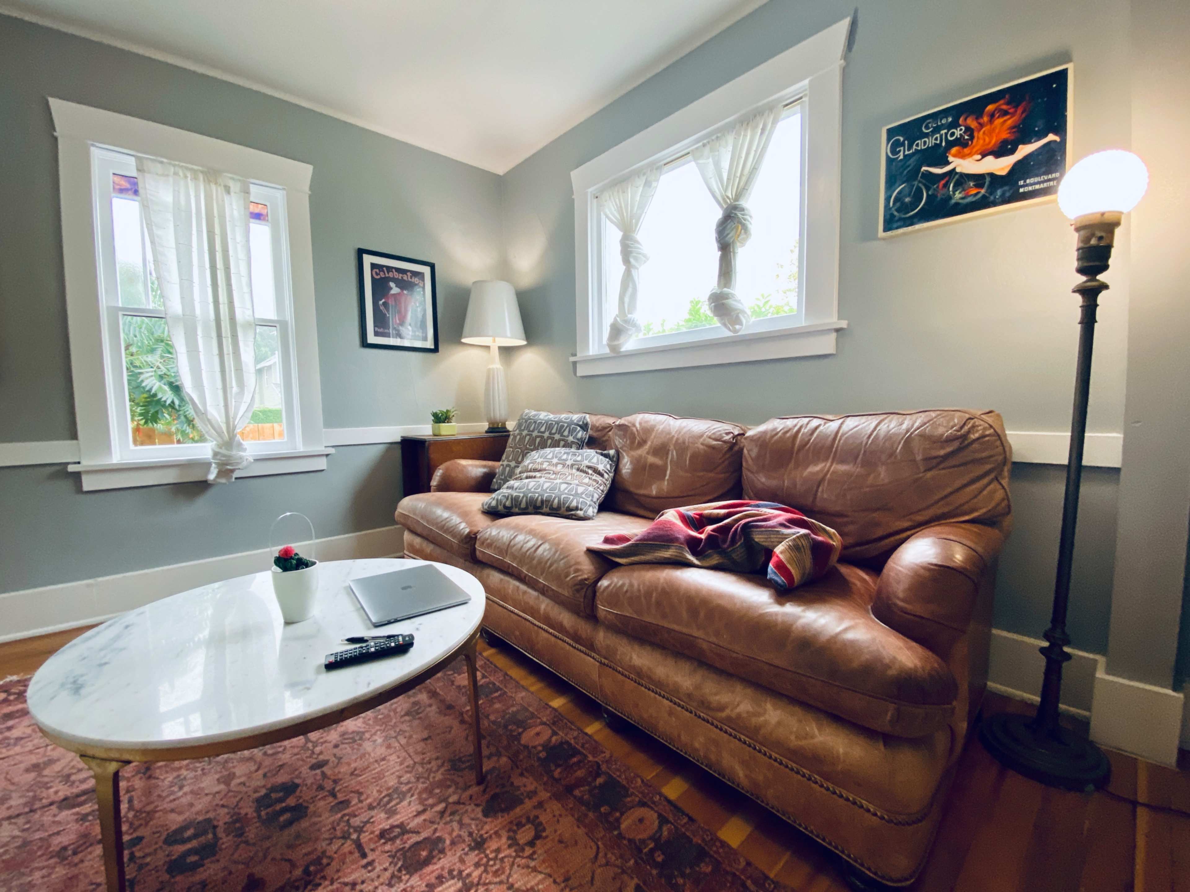 The image shows a cozy living room with a brown leather couch, a marble coffee table, and two large windows letting in natural light.
