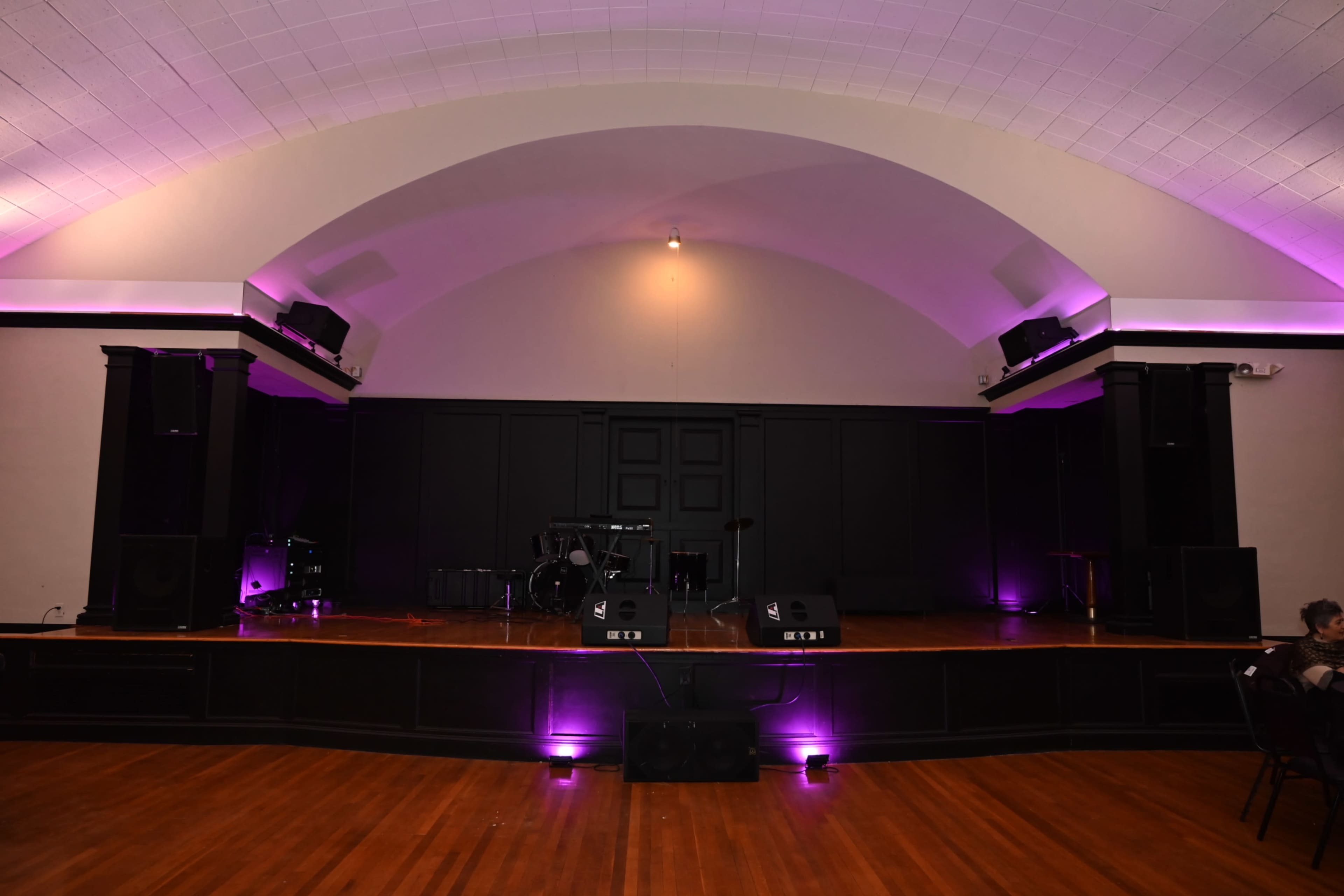 The image shows a music performance stage with a wooden floor, black backdrop, and purple lighting.