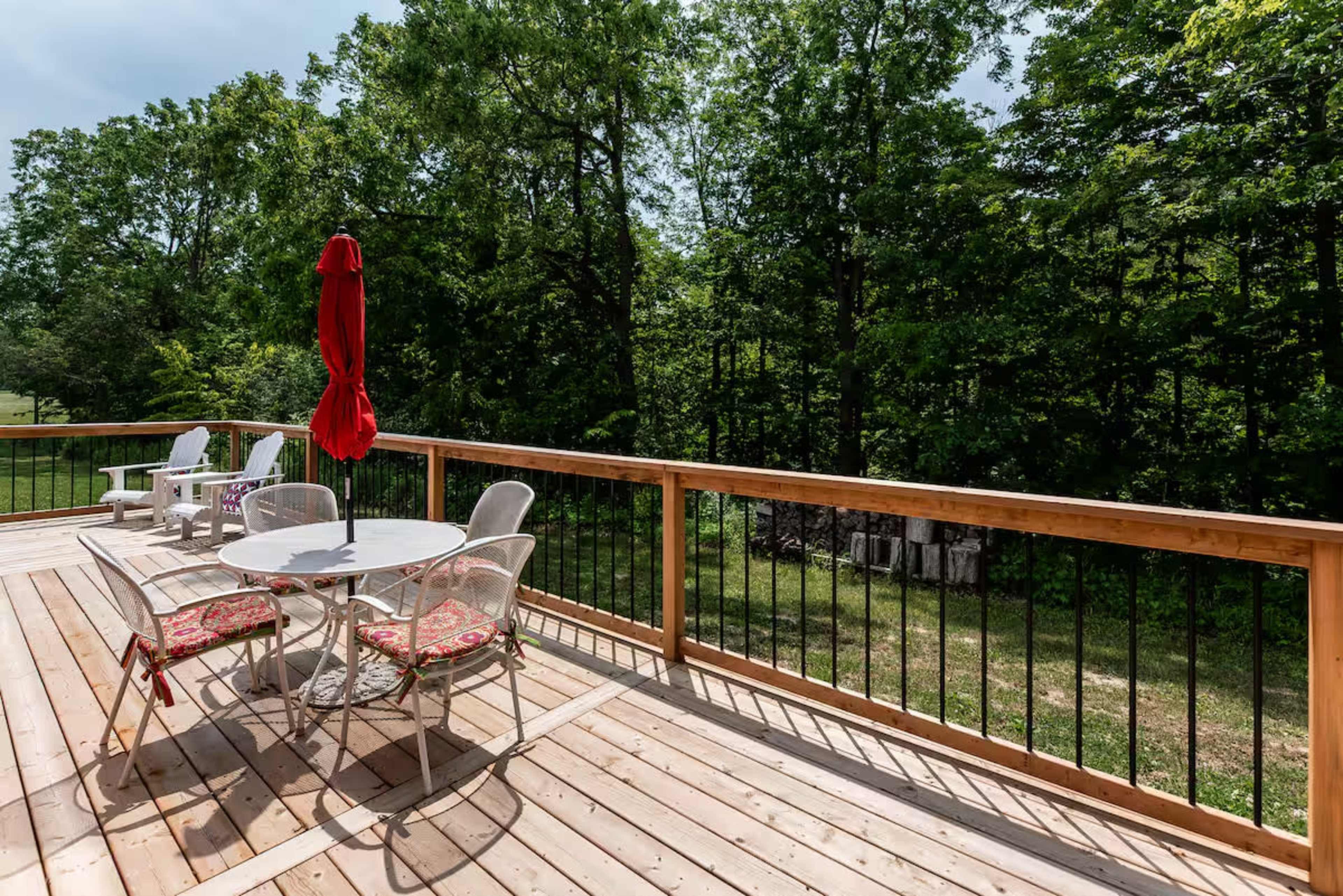 A wooden deck features a round table with chairs and a large red umbrella, surrounded by trees.