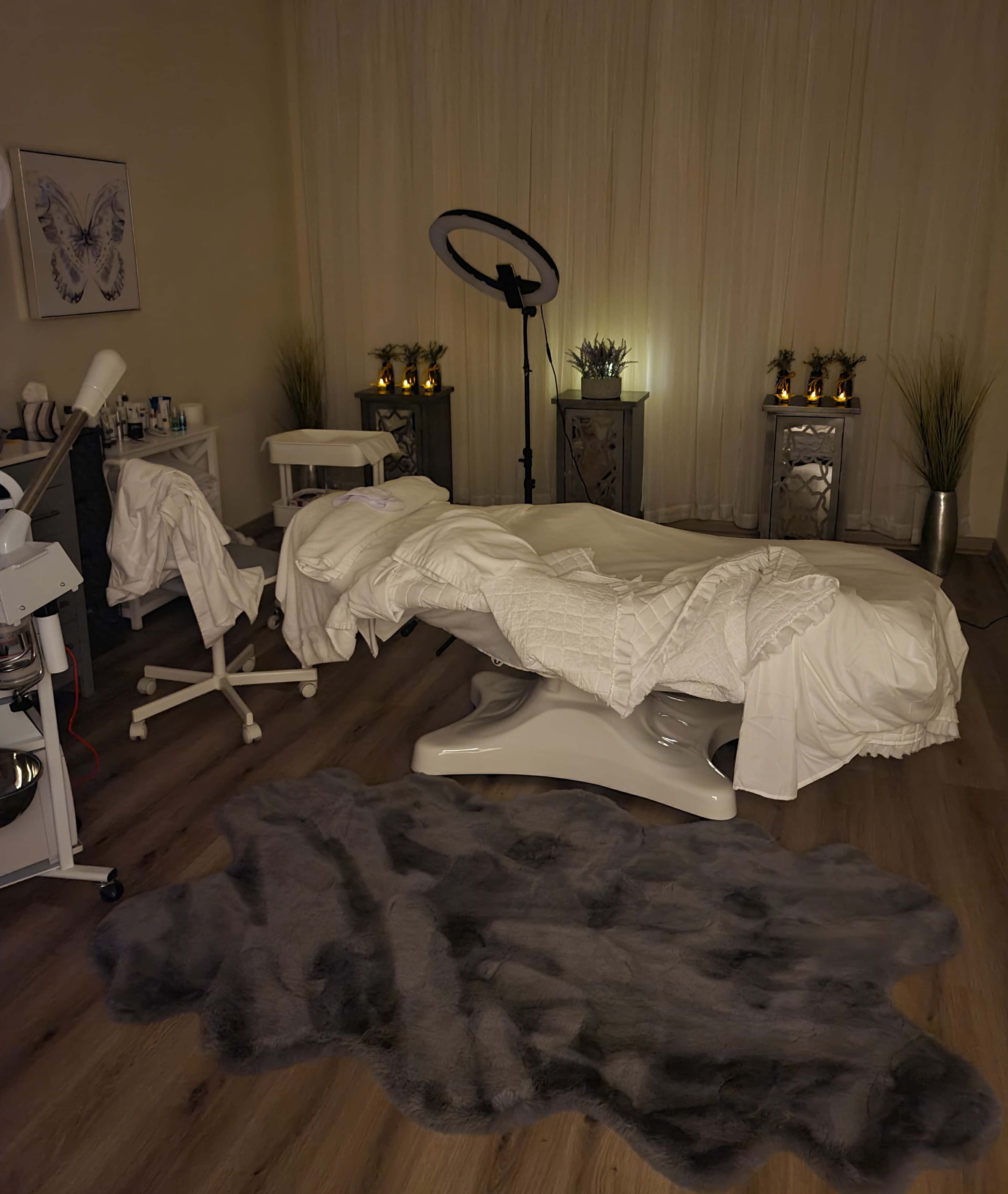 A spa treatment room with a massage table covered in white linens, surrounded by a stool, a floor rug, and softly lit decor elements.