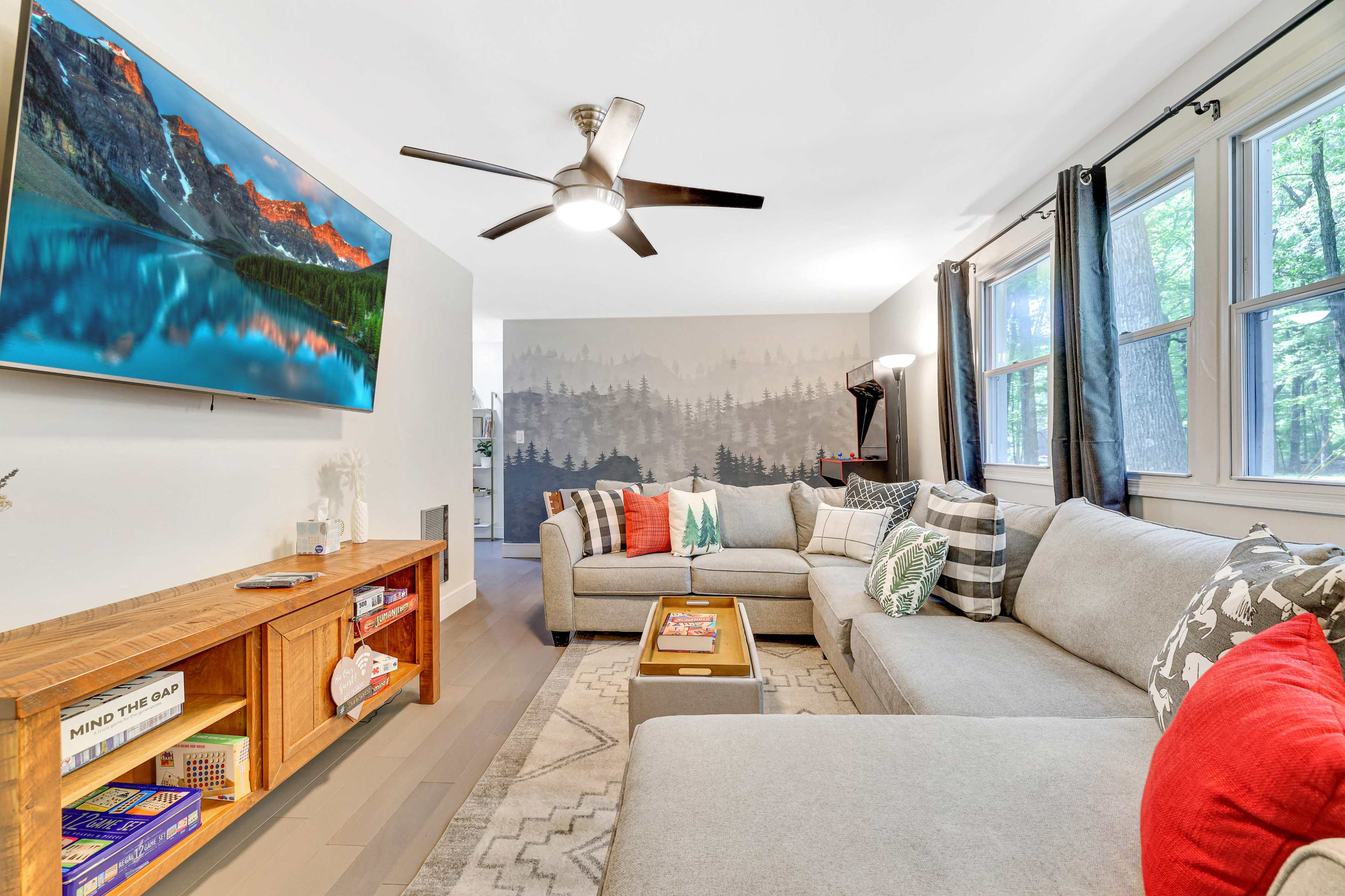 A living room with a sectional sofa, a wooden coffee table, a television on the wall, and large windows allowing natural light to enter.