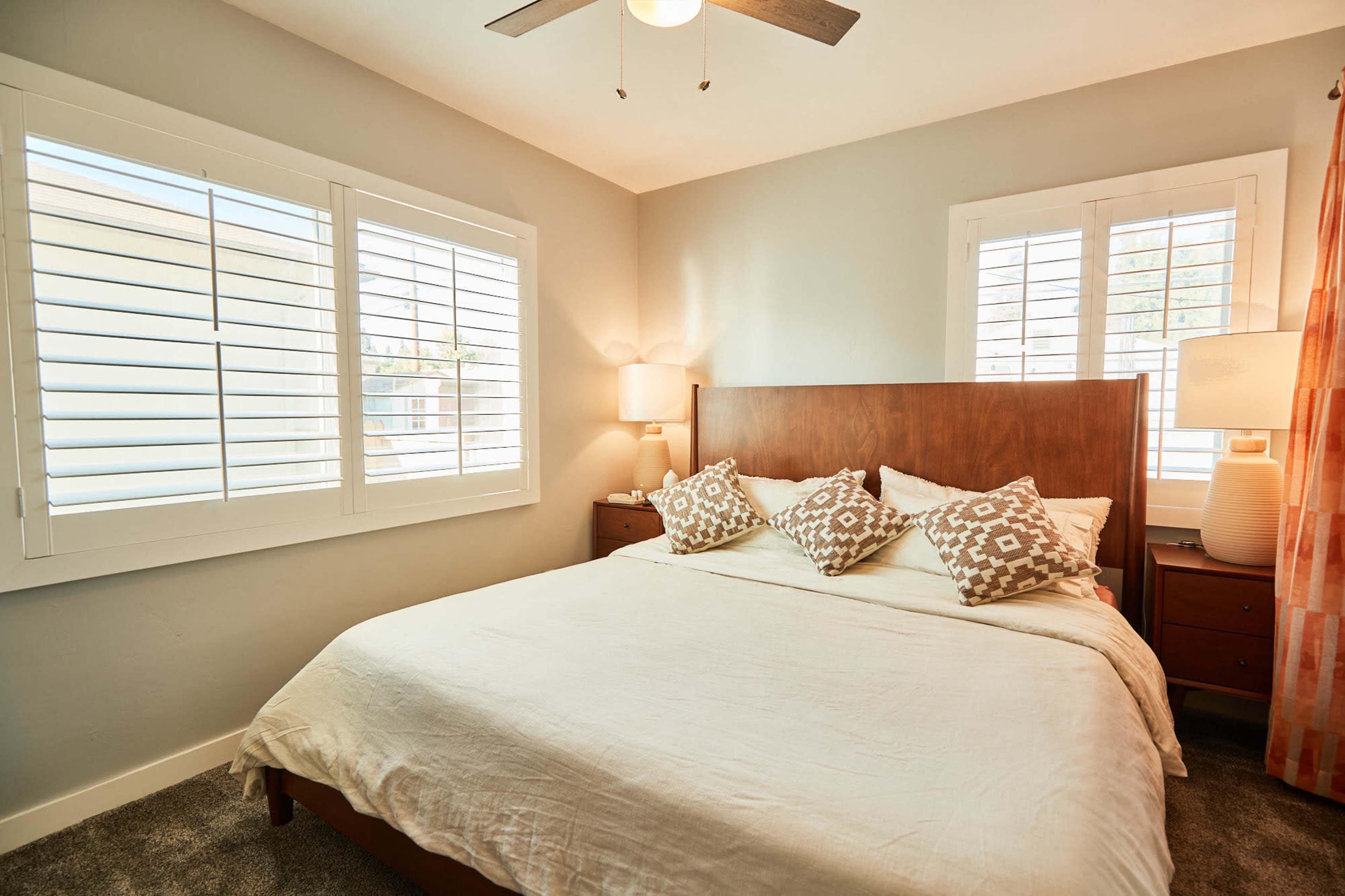 A well-lit bedroom with a wooden bed, decorative pillows, nightstands, and window shutters.