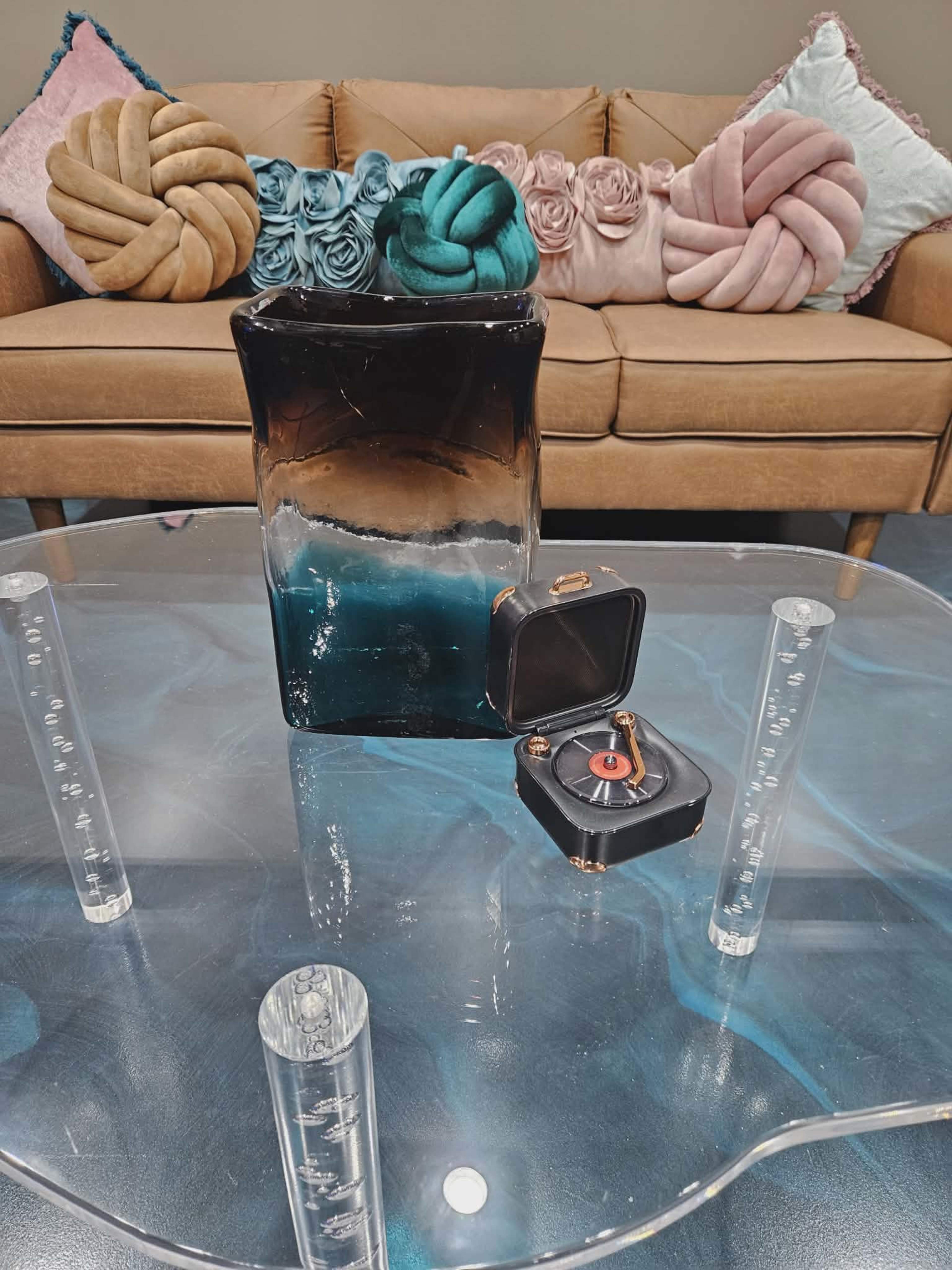 A glass vase with a gradient design sits on a clear table, accompanied by a small round box and transparent cylindrical objects, all in front of a brown sofa adorned with decorative pillows.