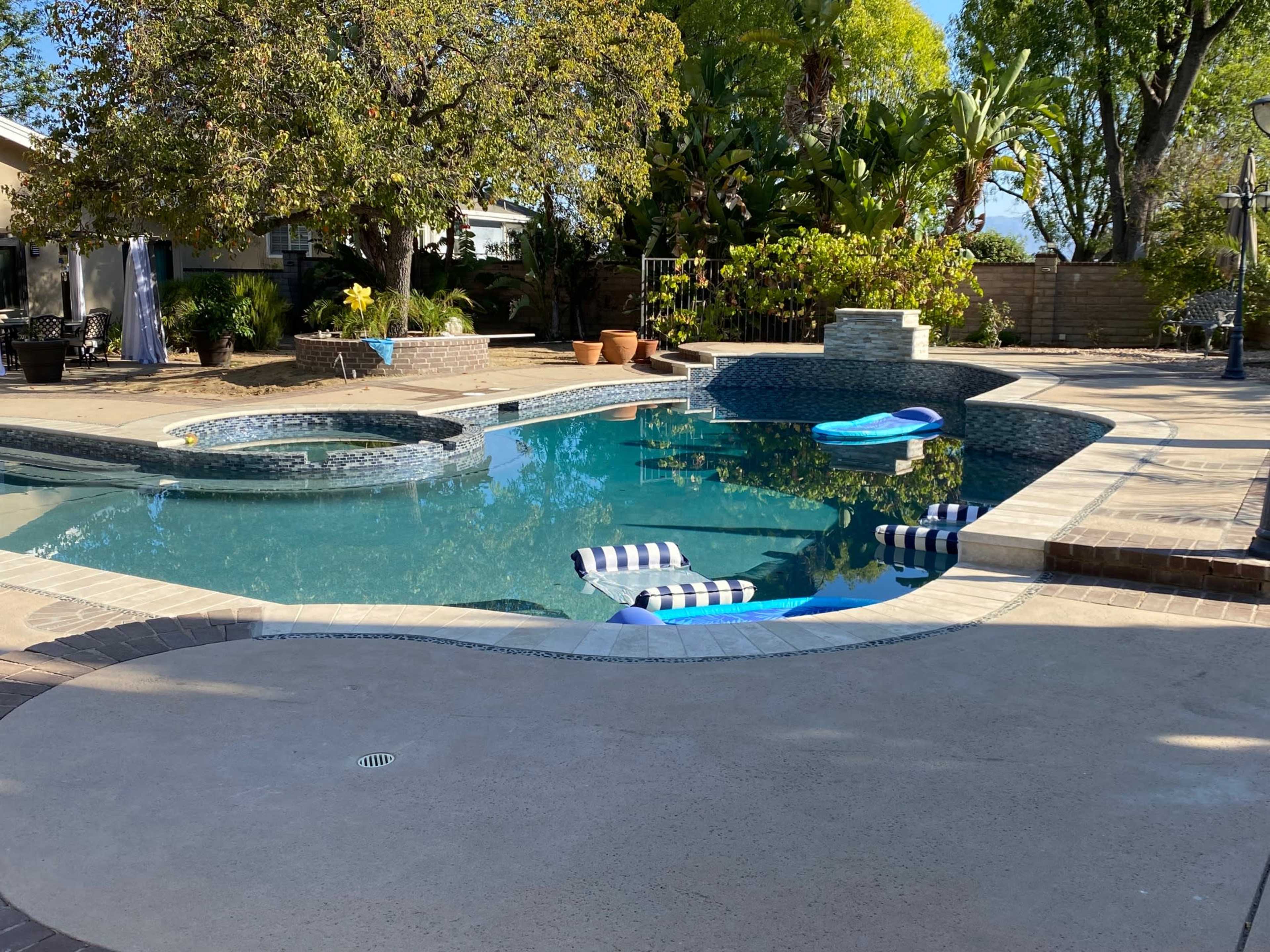 The image shows a swimming pool area with a hot tub, lounge chairs, and surrounding greenery.