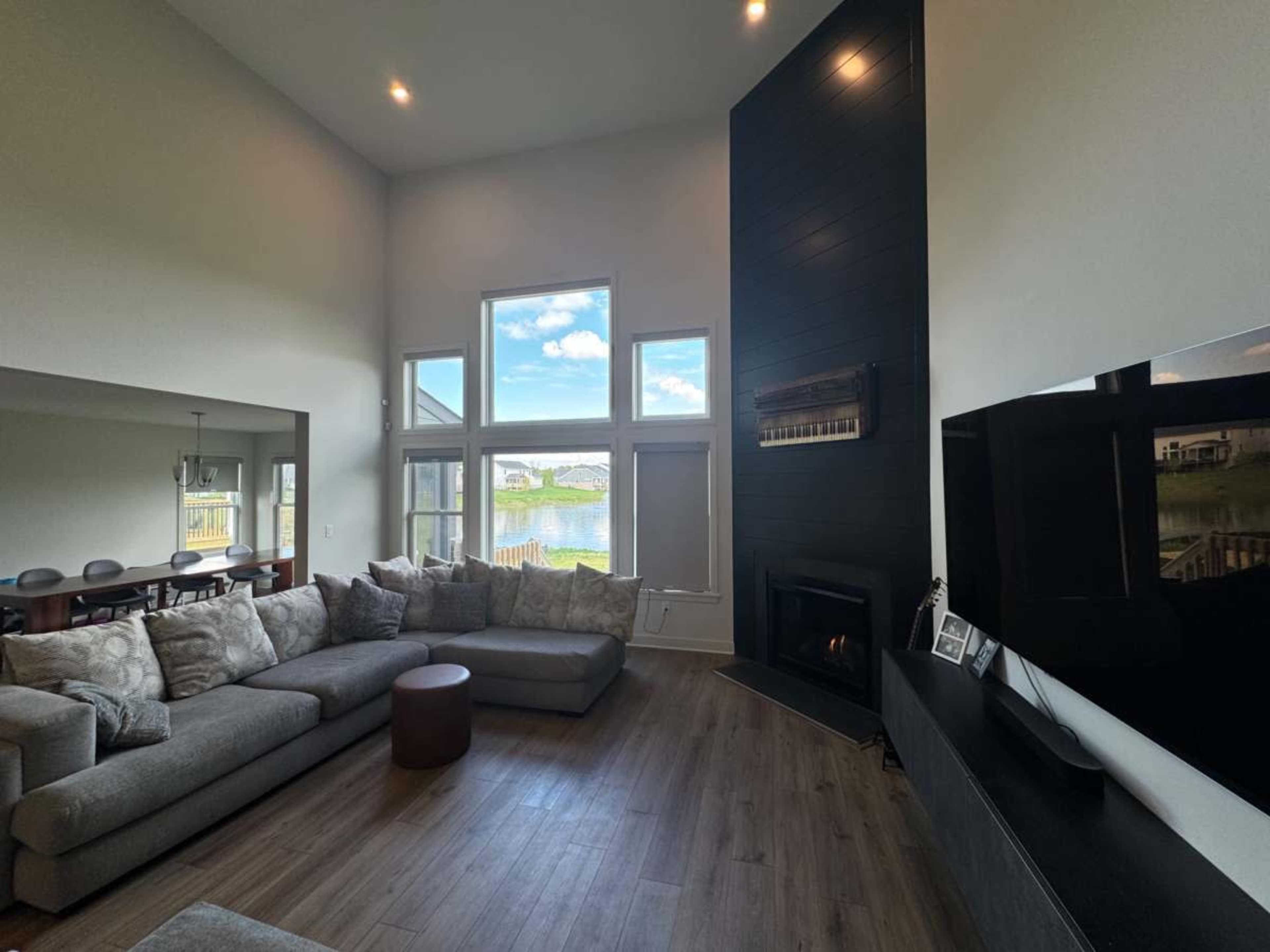 A modern living room features a large sectional sofa, a fireplace, and panoramic windows overlooking a water view.