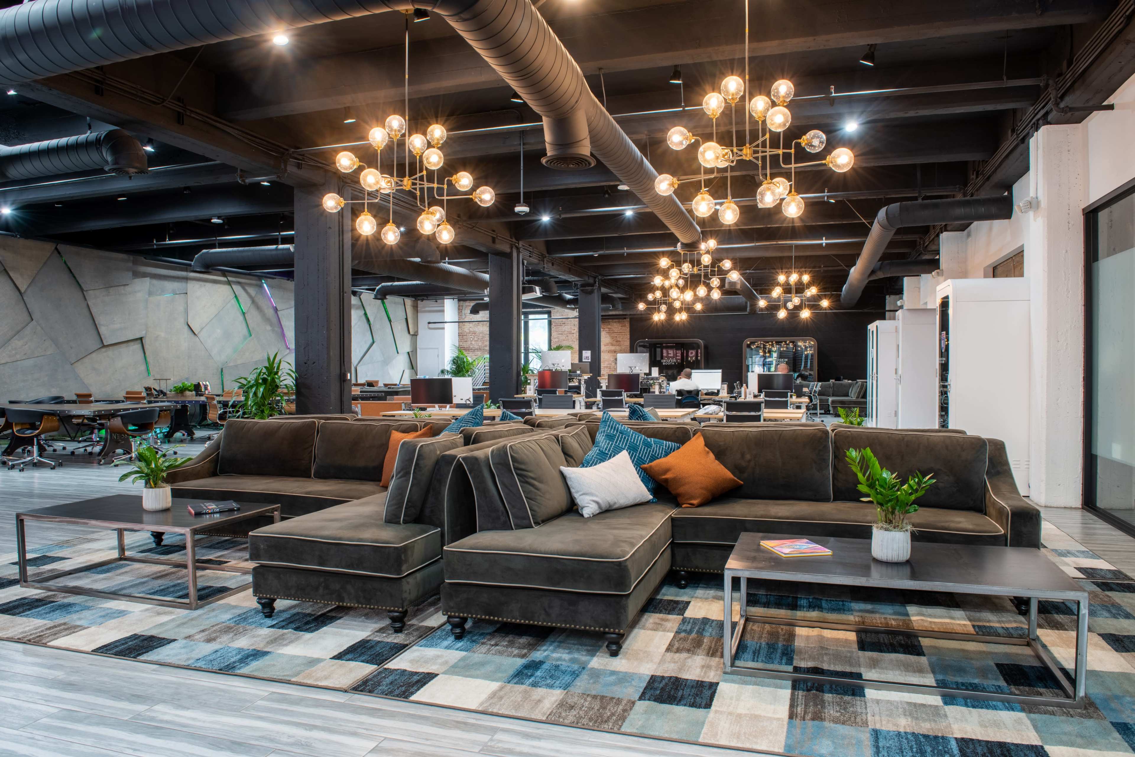 A modern office lounge area featuring a large sectional sofa, decorative lighting fixtures, and plants, with a workspace in the background.