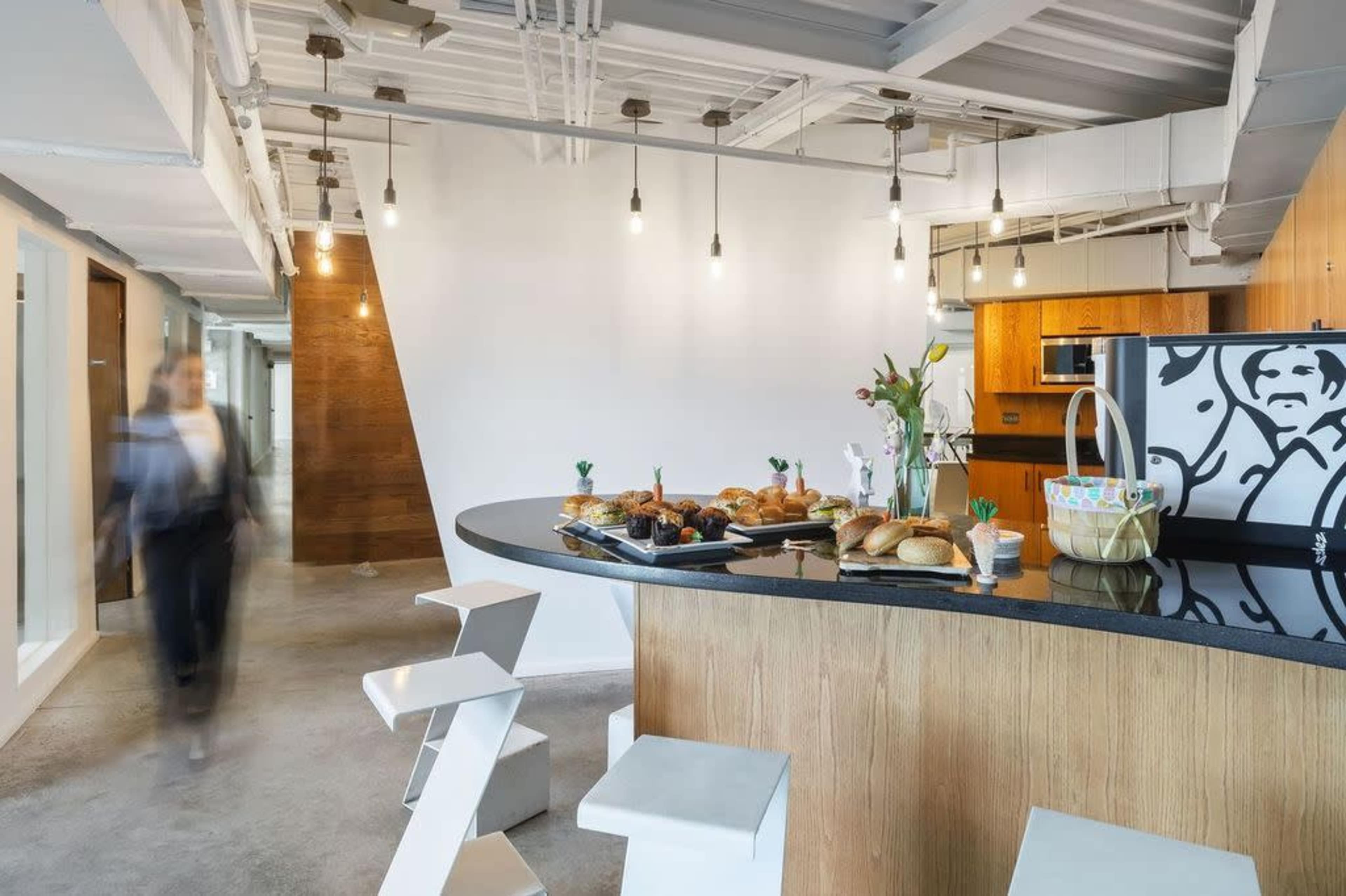 A modern office kitchen features a large black countertop with a variety of pastries, surrounded by a minimalist design and illuminated by hanging light fixtures.