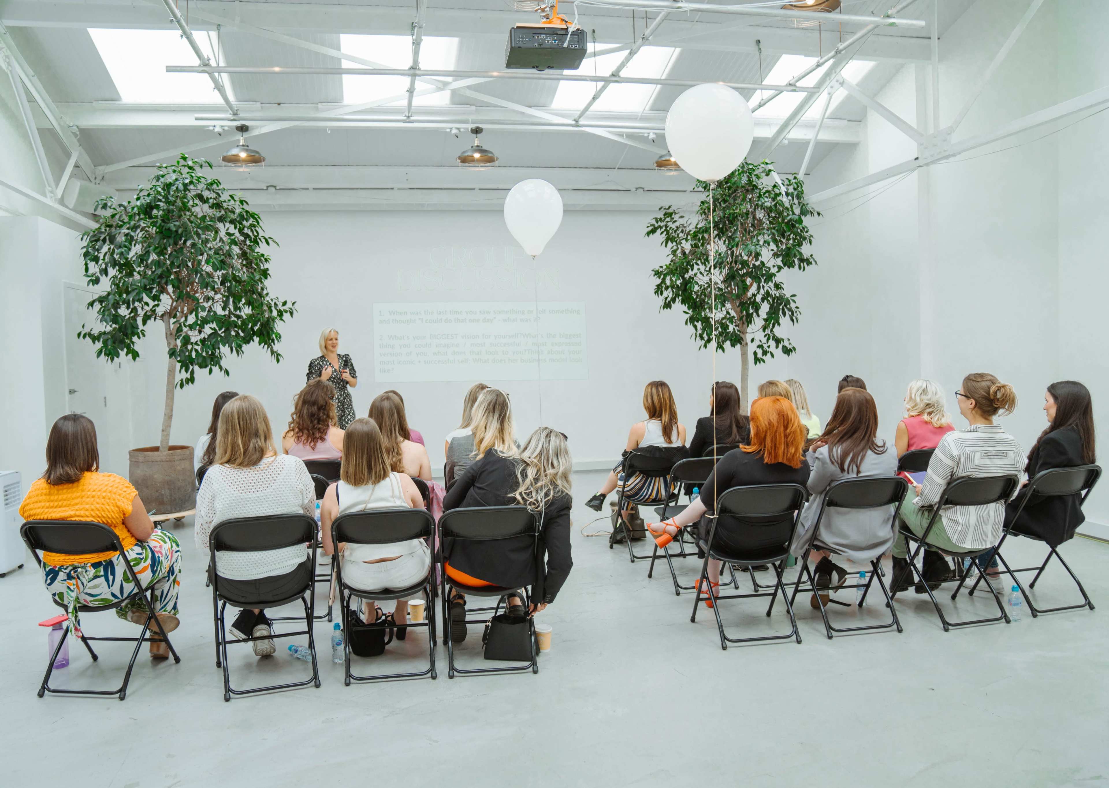 A group of women sits in chairs, facing a speaker in a spacious, light-filled room with two large potted plants and a projected presentation on the wall.