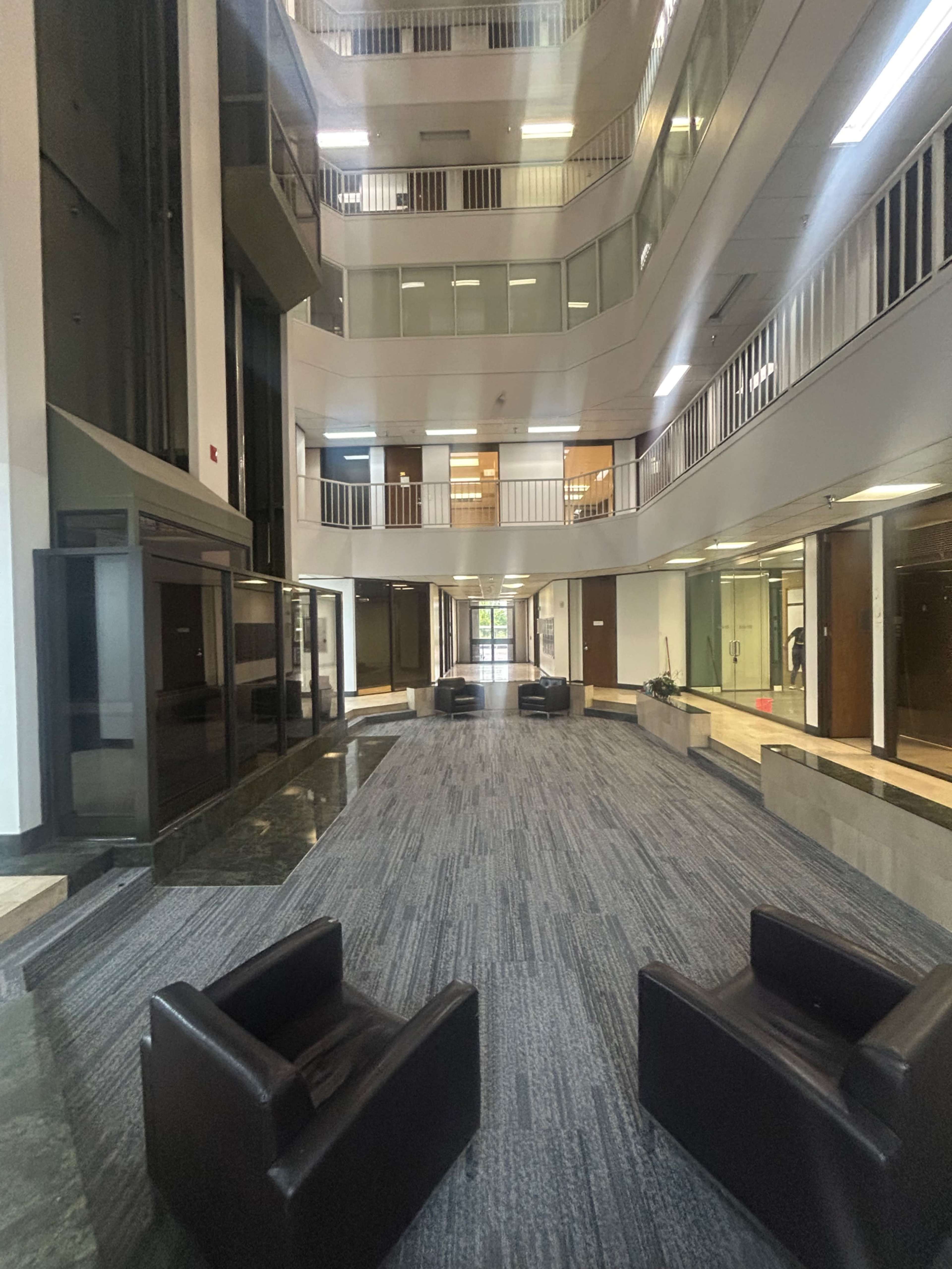 The image shows a modern, spacious atrium with multiple levels, featuring leather chairs arranged on a patterned carpet and large glass windows.