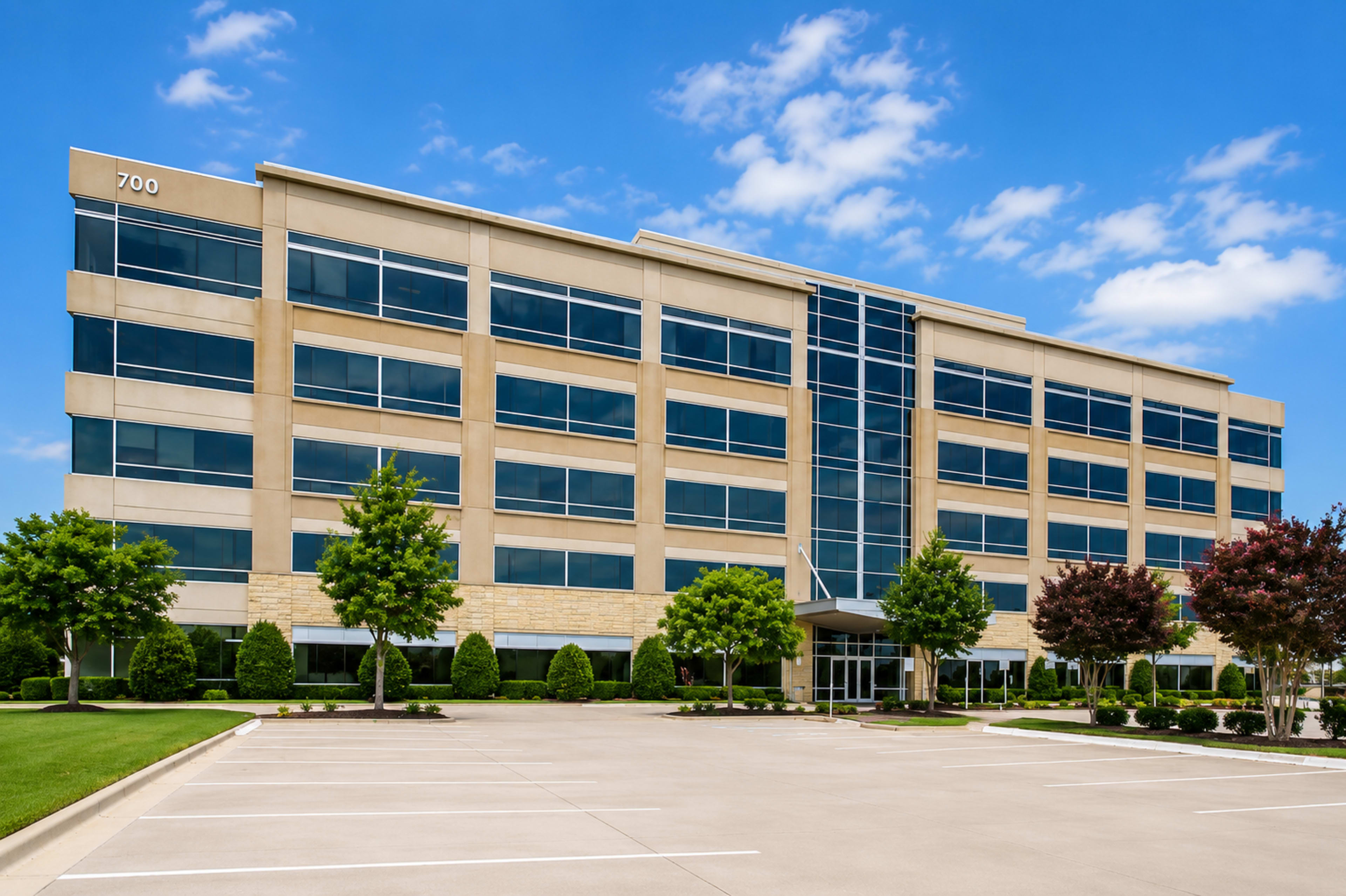 The image shows a modern, multi-story office building with large windows and landscaped parking in front.